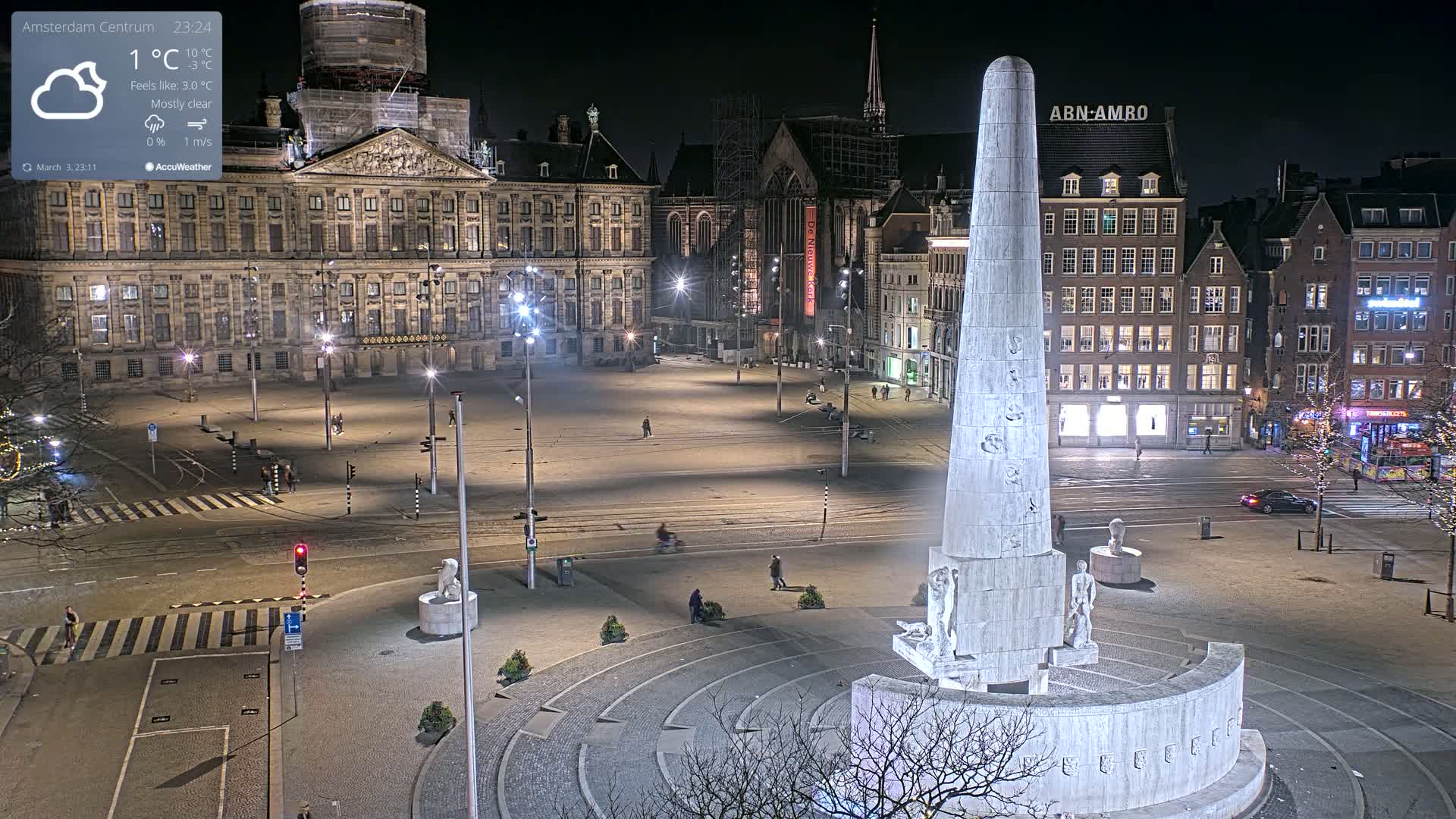 A mostly clear night scene of a large, open square in a city, centered around a tall, light-colored monument, with historical buildings and sparse pedestrian traffic.