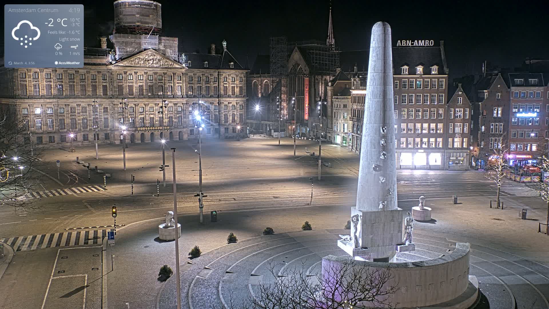 A mostly empty, snow-dusted city square at night is illuminated by streetlights, featuring a large monument at its center and a palace in the background under light snow.