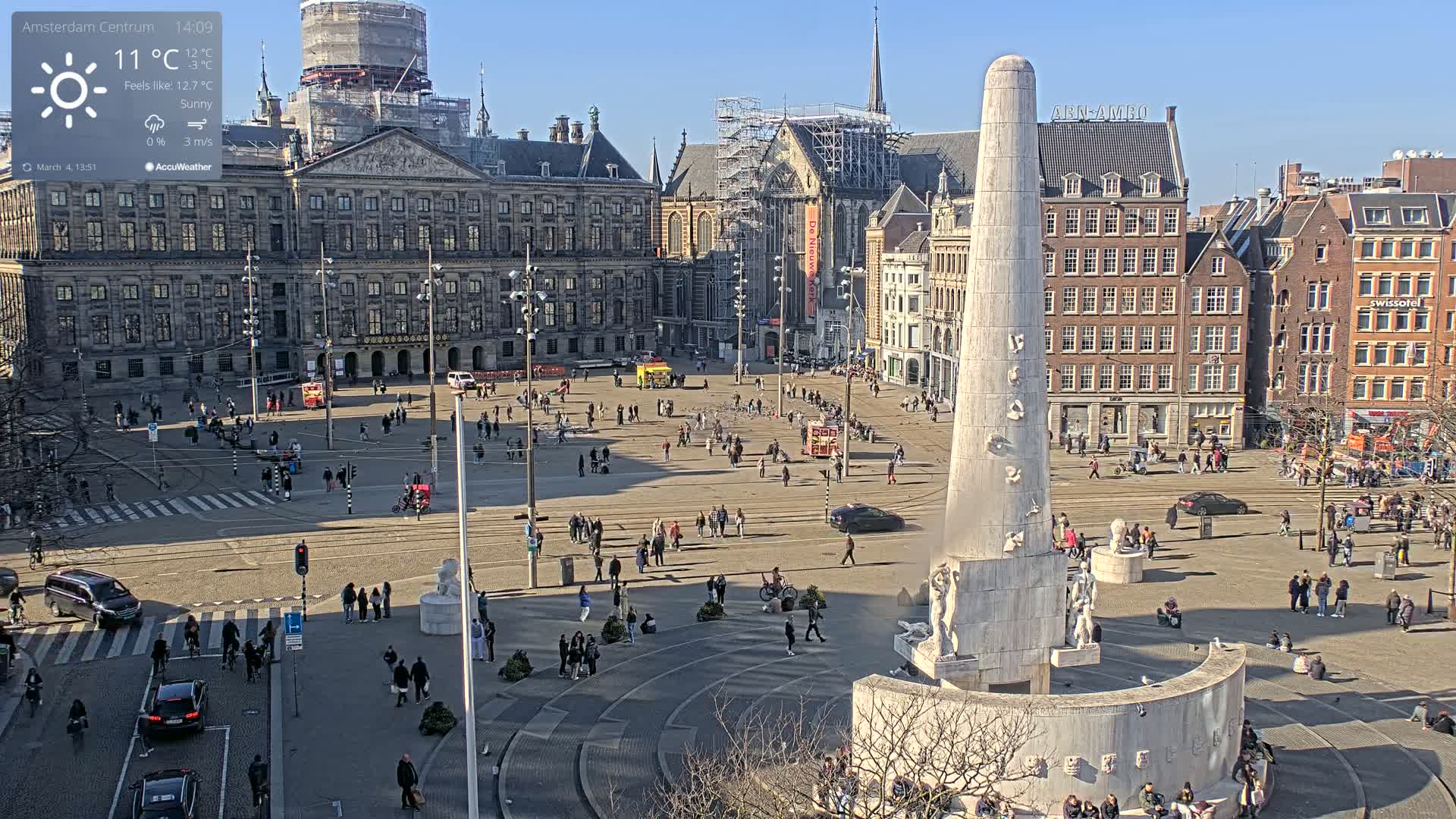 A sunny day in a large city square with a tall monument at the center, surrounded by historical buildings and people walking around.