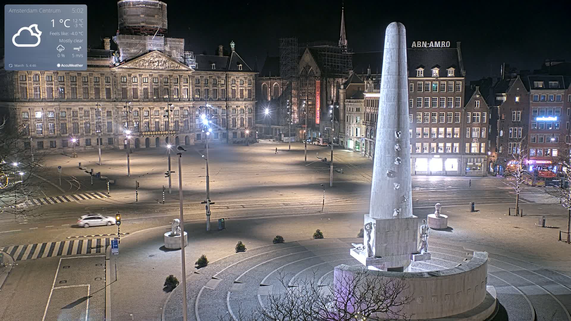 A mostly clear night scene shows a large, empty square at night, centered on a tall, light-colored monument with figures at its base, with buildings and streetlights illuminating the area.