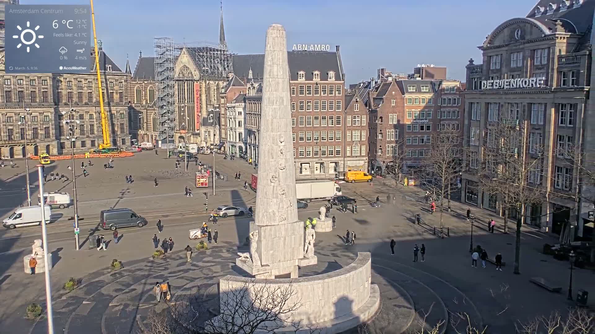 A sunny city square features a tall monument, surrounded by buildings and pedestrians, under a clear blue sky.