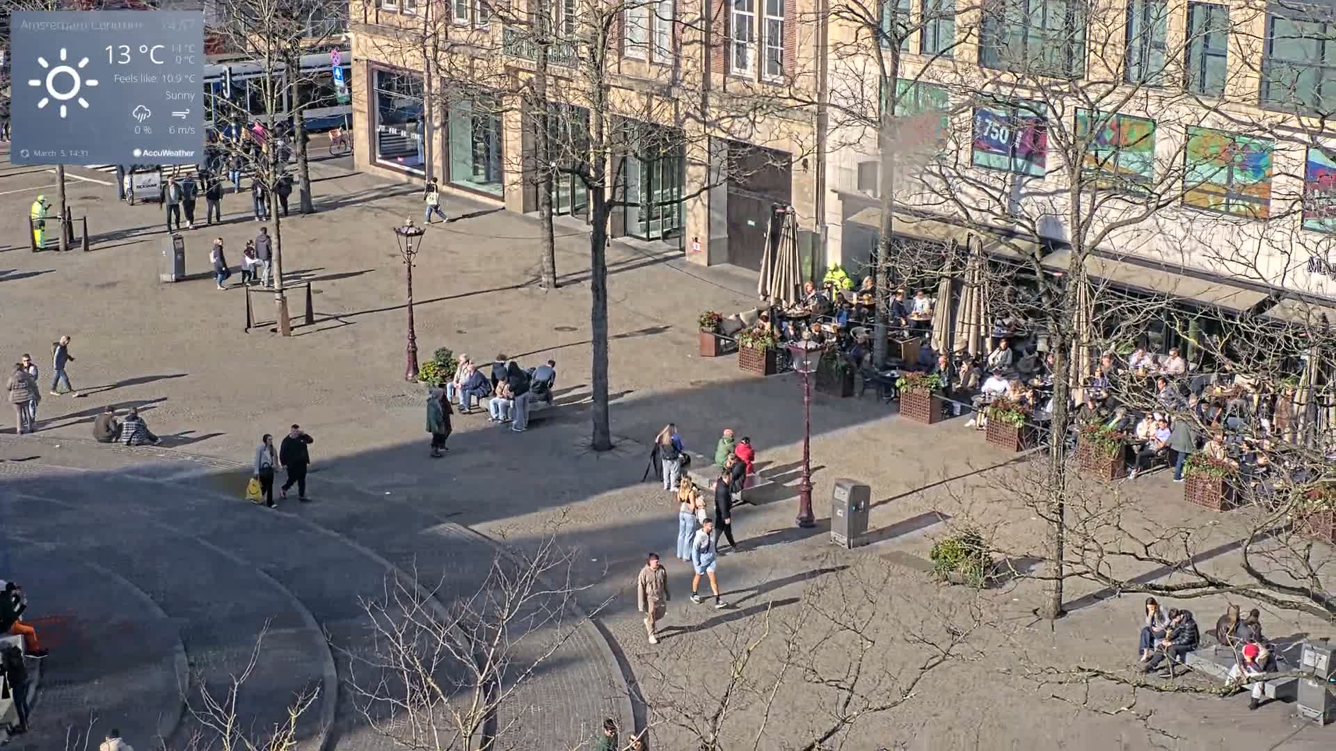 A sunny plaza with many people walking and sitting at outdoor cafes, surrounded by buildings with bare trees.