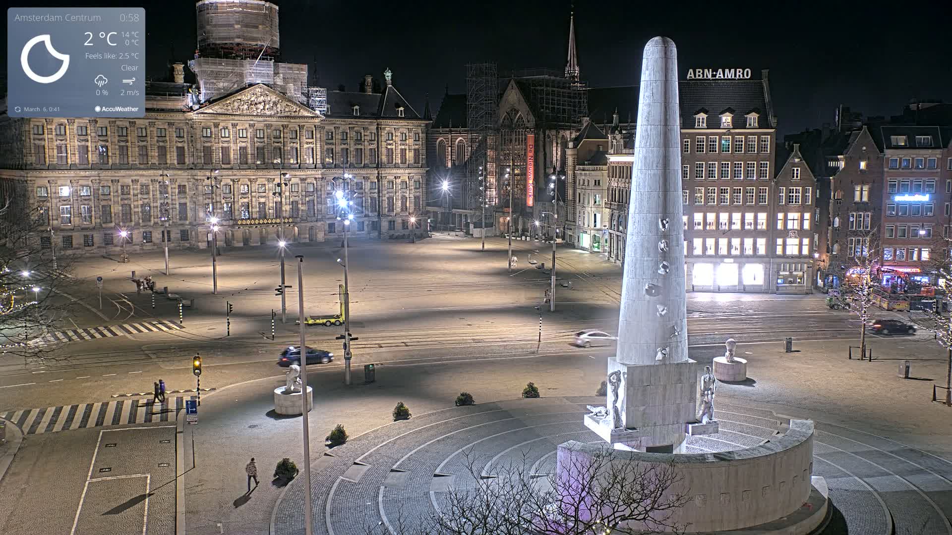 A nighttime, clear view of a city square, centered on a tall, stone monument, with a palace and other buildings illuminated in the background.