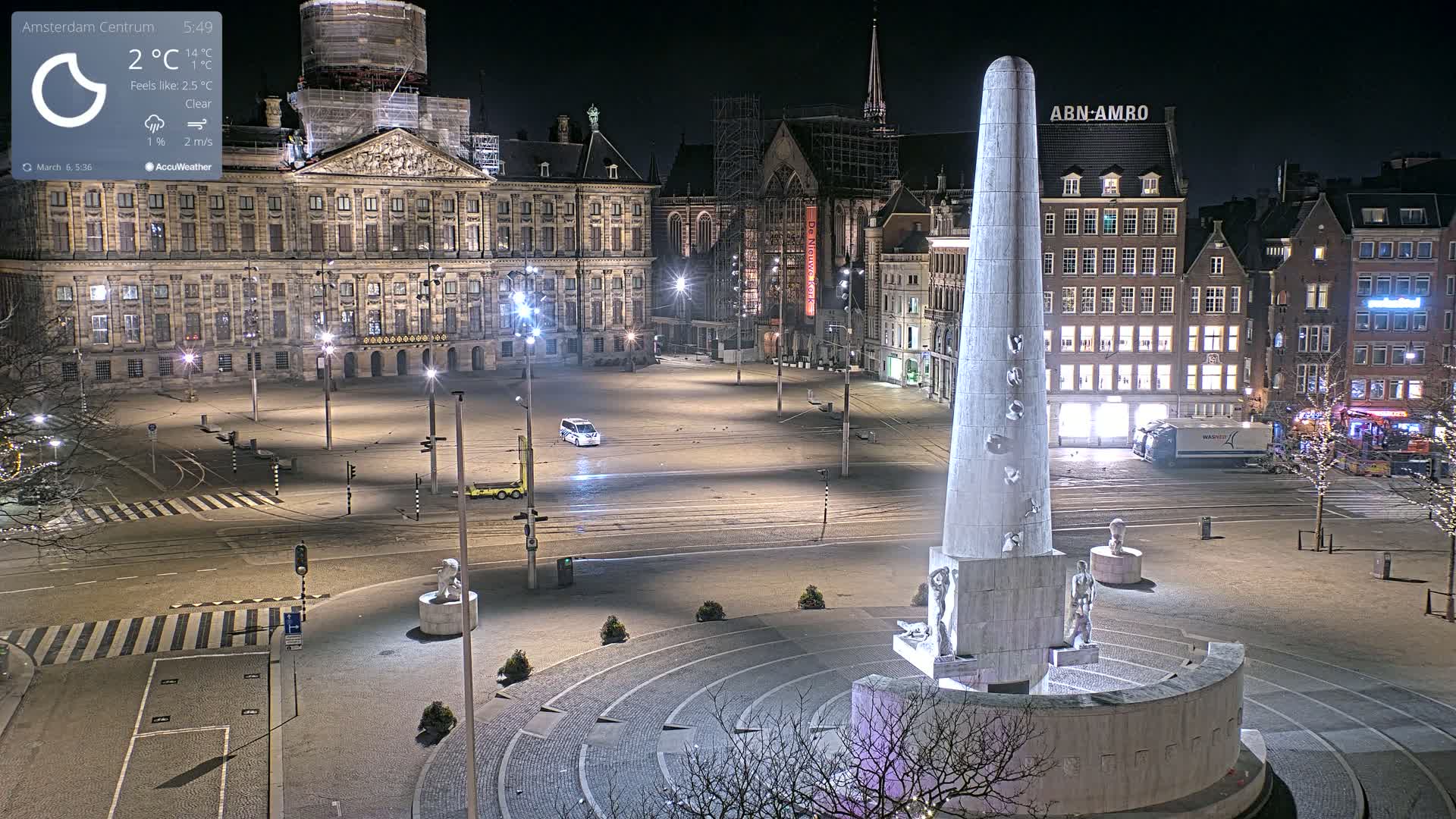 A mostly empty city square at night, illuminated by streetlights, features a tall monument in the foreground and a large palace in the background under clear skies.