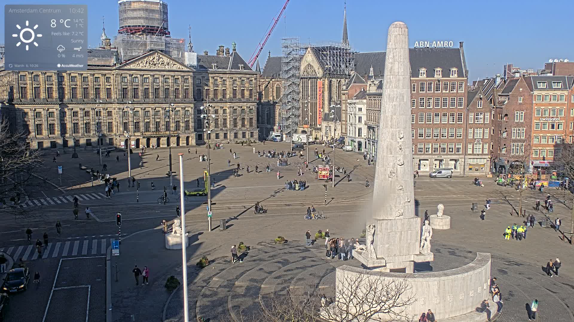 A sunny city square, with a large monument in the center, surrounded by historical buildings, one of which is under construction.
