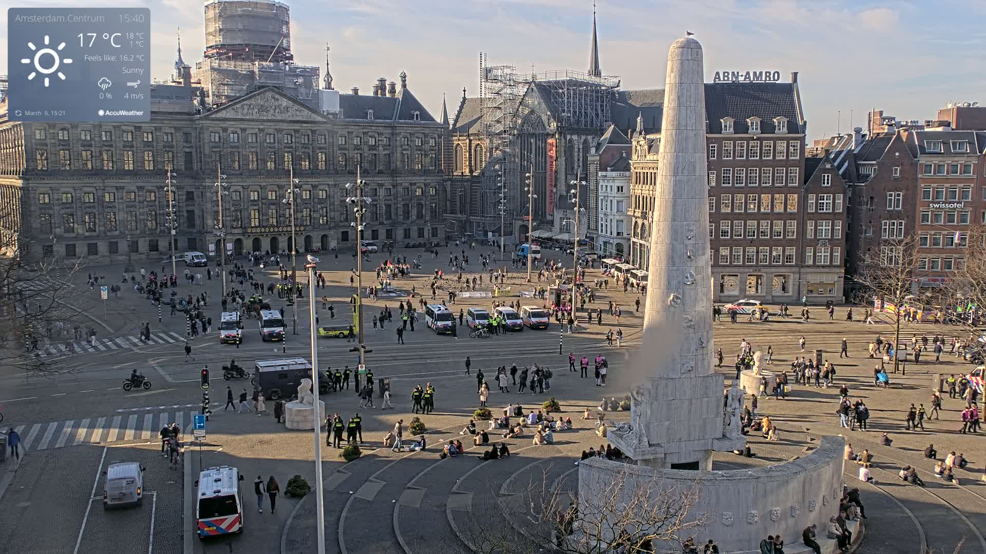 A sunny city square with a large monument at its center, surrounded by numerous people and several police vehicles.