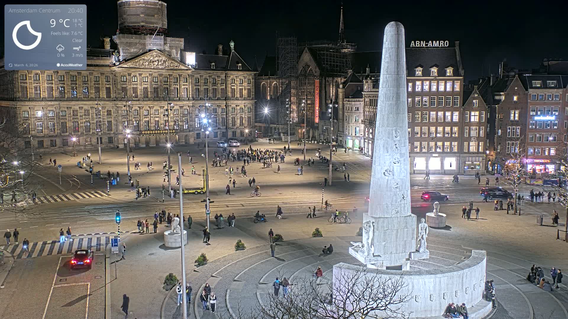 A nighttime, clear-weather view of a large city square with a tall monument at its center, surrounded by buildings and people.
