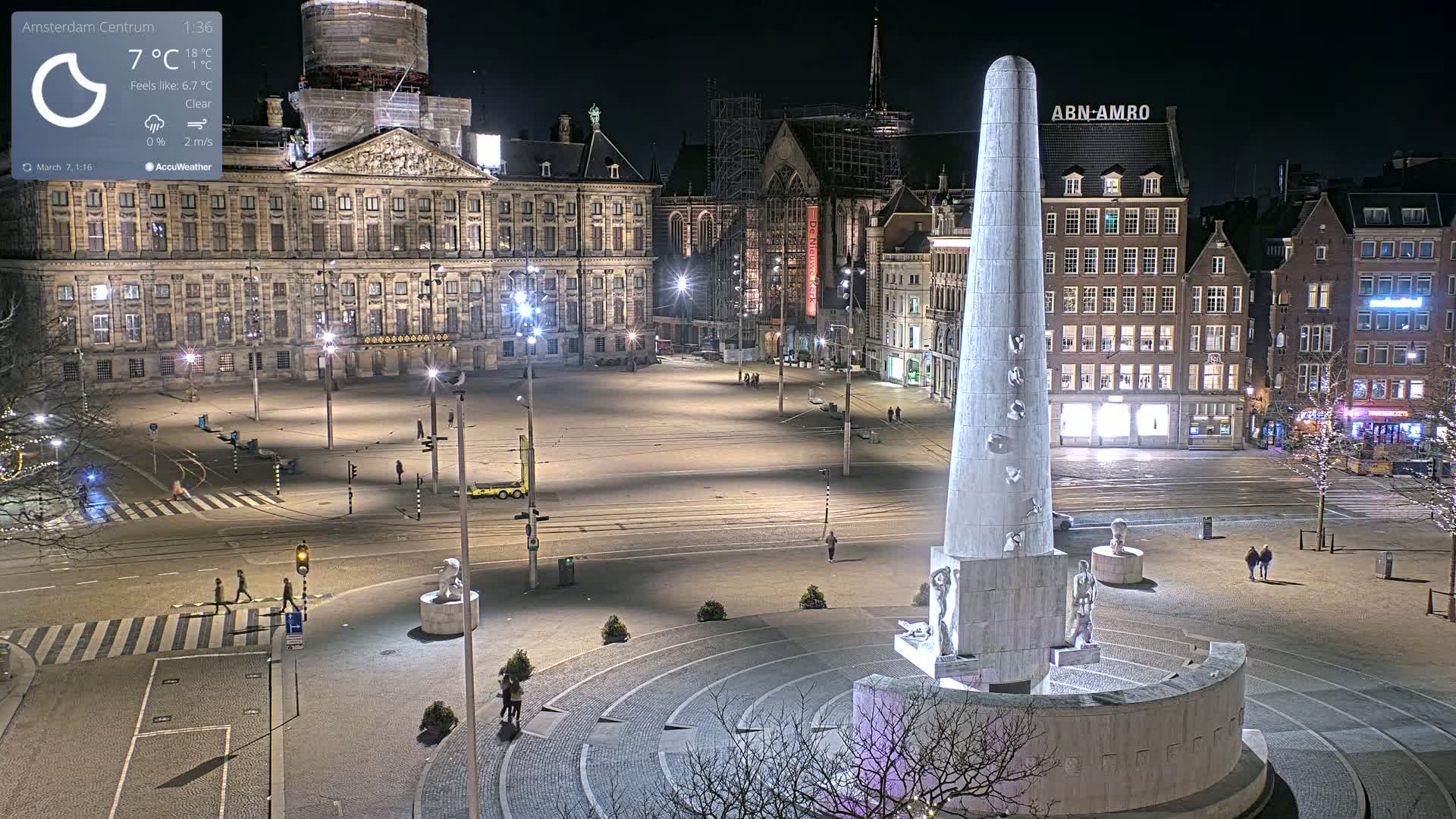 A nighttime, clear view of a large city square with a tall monument at its center, surrounded by historic buildings and sparsely populated with pedestrians.