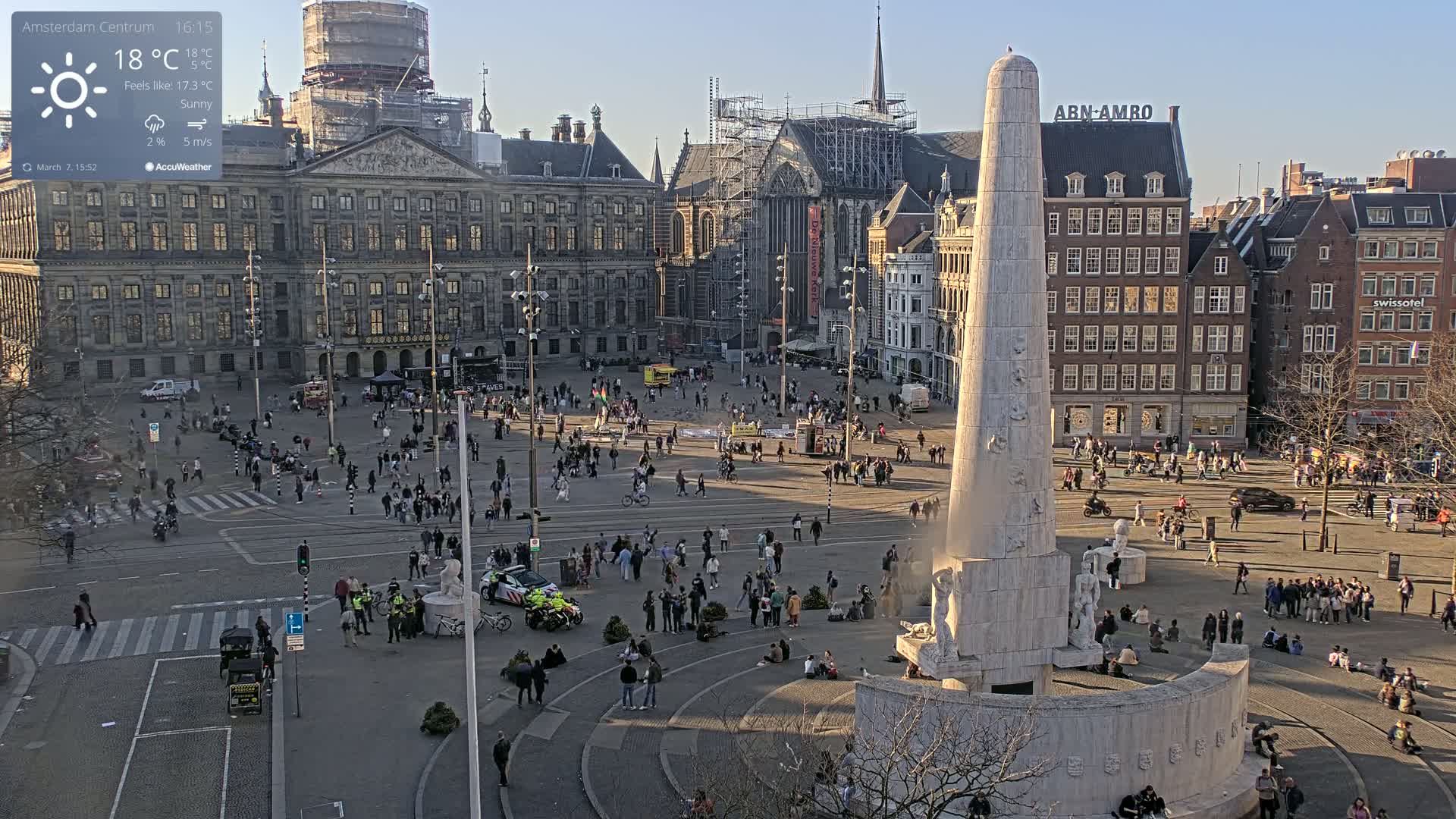 A sunny city square with a large monument at its center, surrounded by numerous buildings, and many people walking and sitting about.