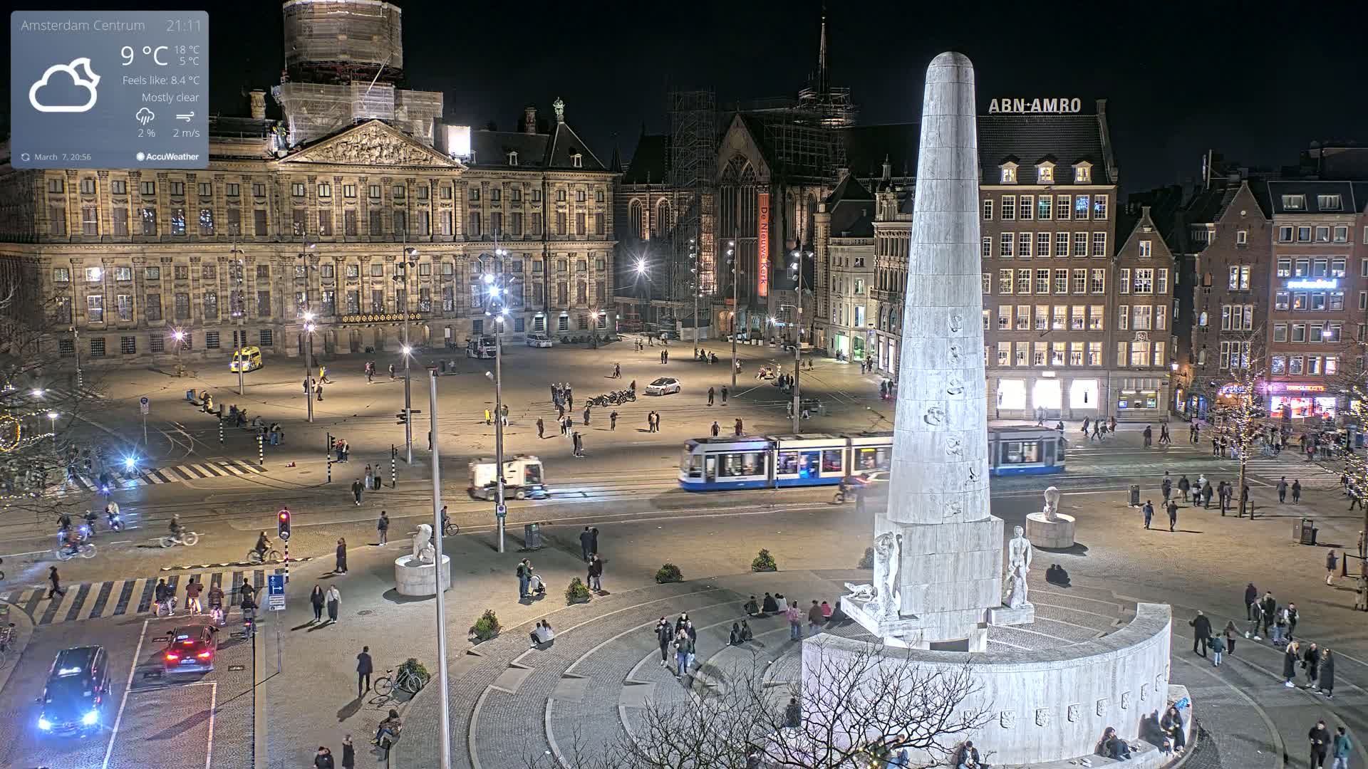 A nighttime view of a city square with a large monument at its center, a tram passing by, and people walking around under mostly clear skies.