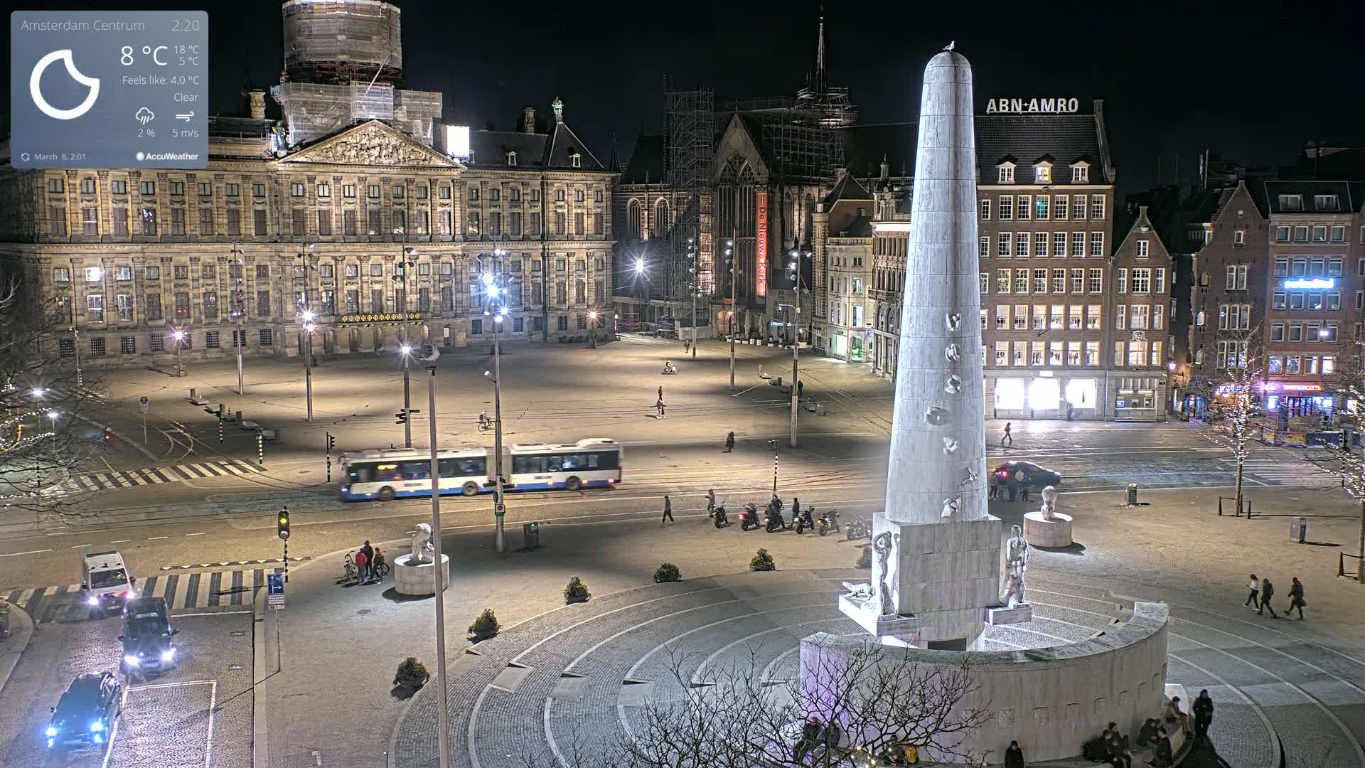 A nighttime view of a large, illuminated city square with a tall monument in the center, several vehicles, and sparse pedestrian traffic under clear skies.