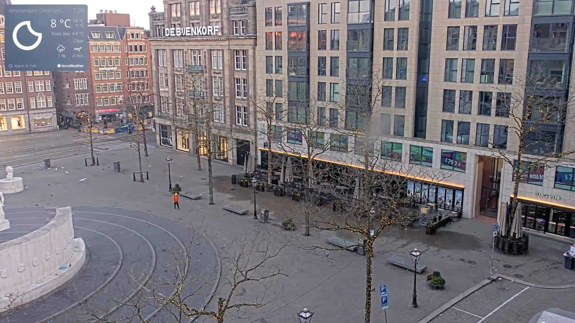 A mostly empty, paved square in a city, under a clear sky, with buildings lining the perimeter, bare trees decorated with lights, and a few people visible.