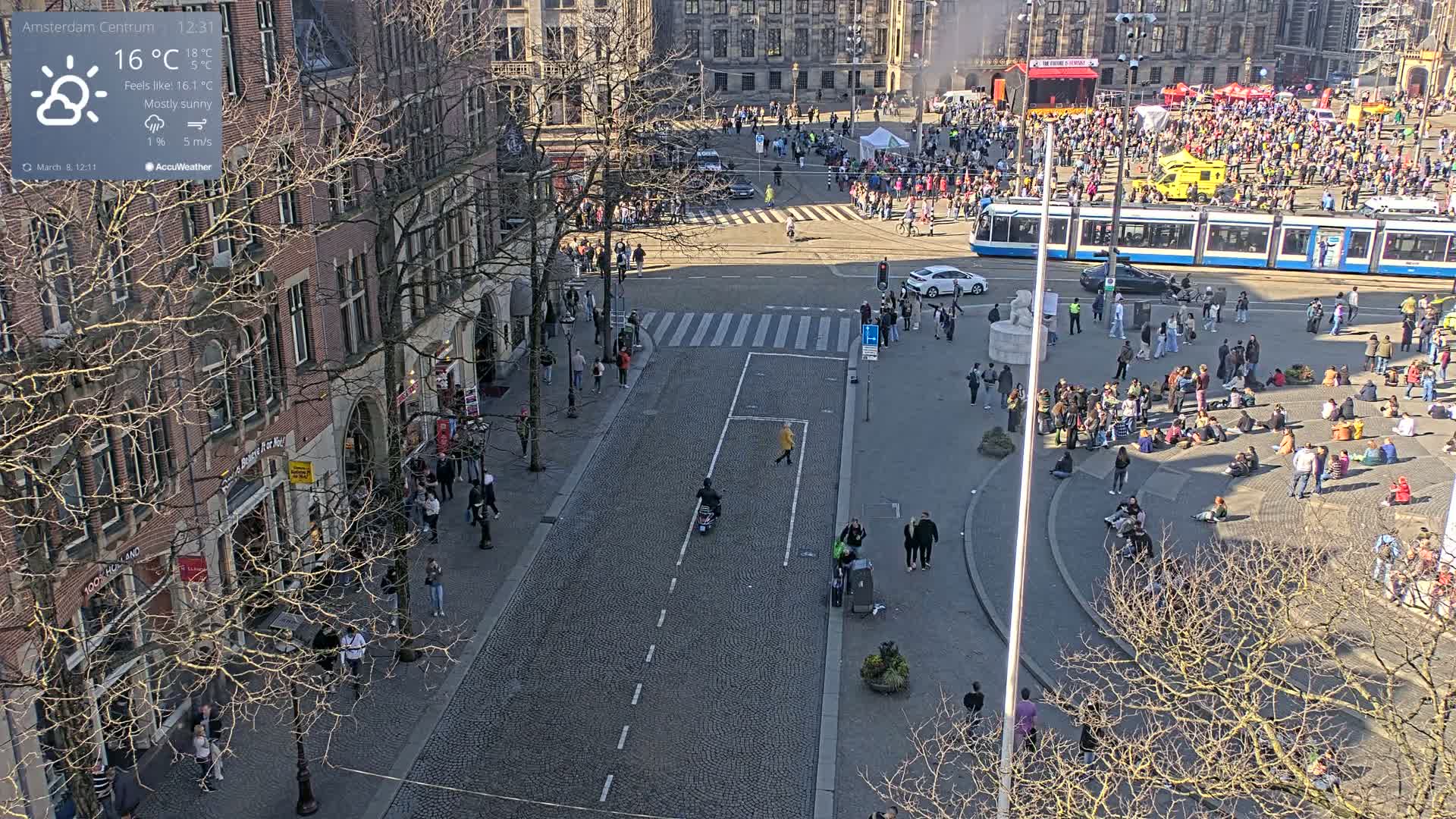 A mostly sunny city square with a crowd of people, some sitting and others walking, and a tram passing by, all viewed from above.