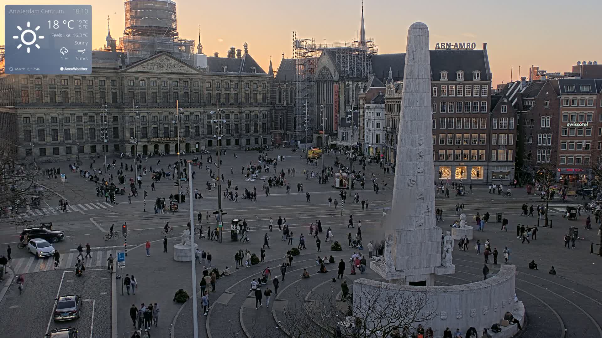 A large, sunlit city square with a monument at its center, surrounded by historic buildings and many people, under sunny skies.