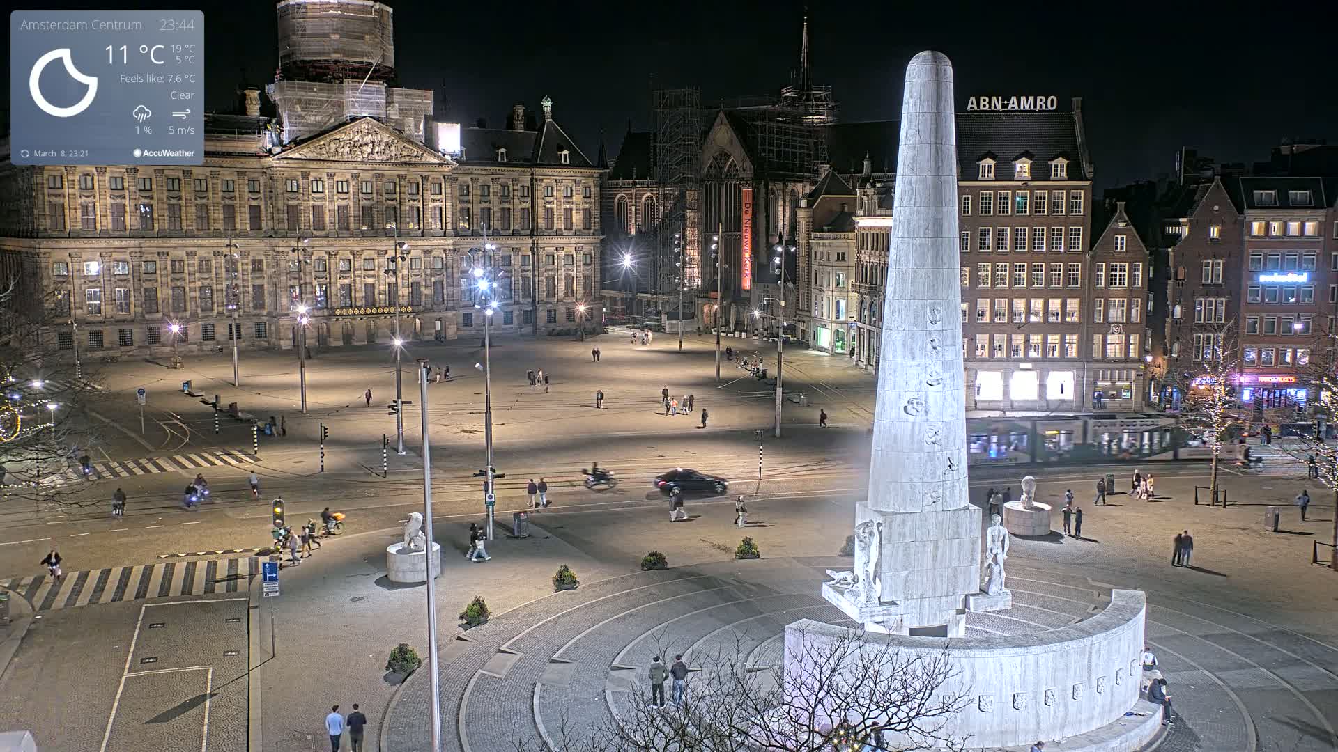 A nighttime, clear view of a large city square with a tall monument at its center, surrounded by buildings and a few pedestrians.