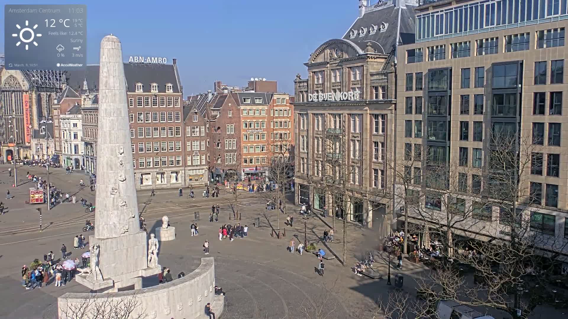 A sunny city square with a tall monument in the center, surrounded by several multi-story buildings and people walking around.