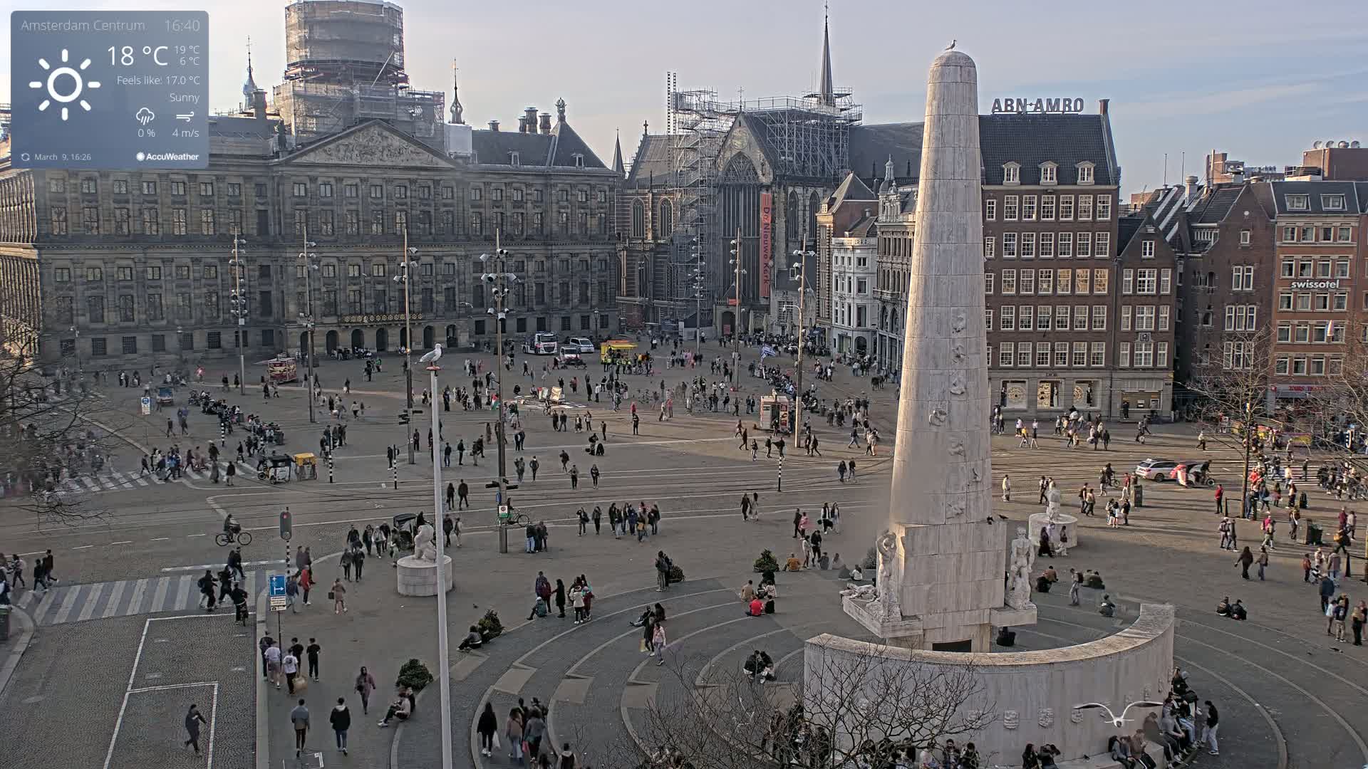 A sunny city square with a tall monument in the center, surrounded by historic buildings and many people walking and sitting.