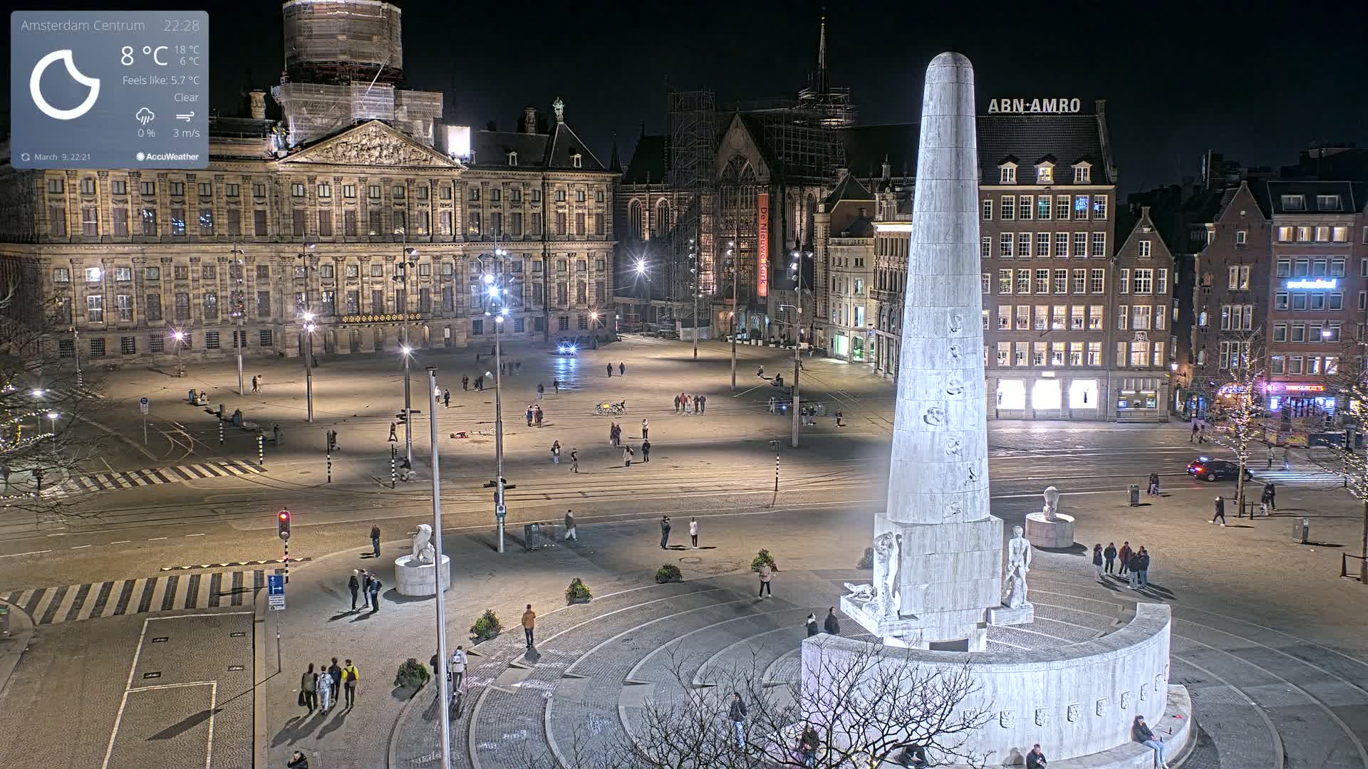 A nighttime, clear-weather view of a large city square with a tall monument at its center, surrounded by historical-looking buildings and a few pedestrians.
