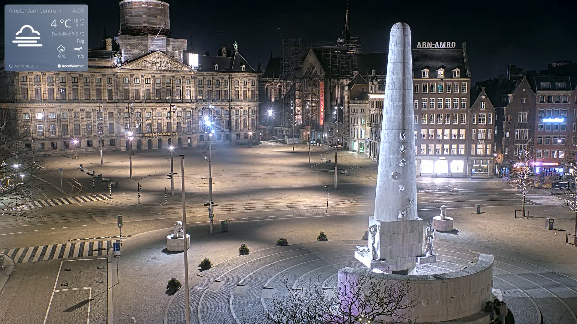 A nighttime, foggy view of a large, empty square with a tall monument in the center, surrounded by illuminated buildings.