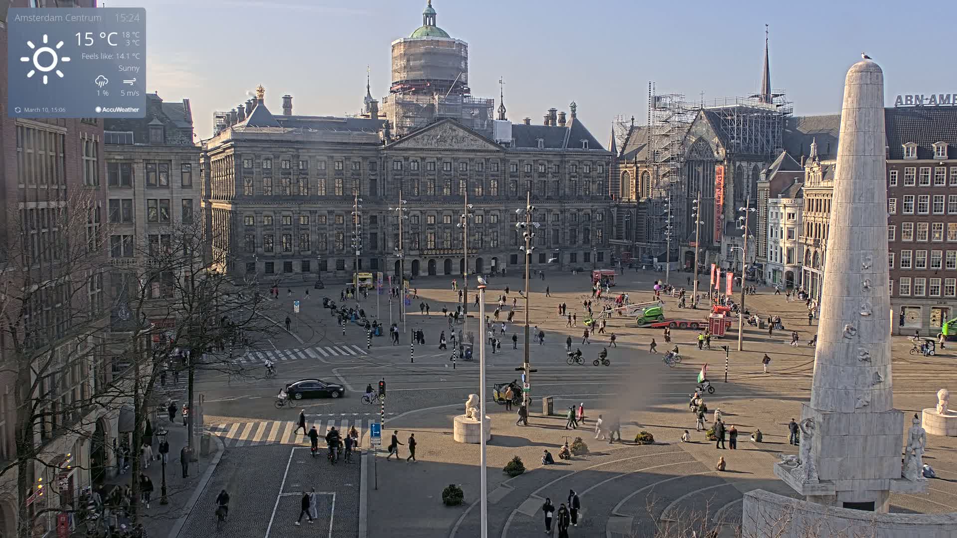 A sunny day in a large city square, showing a palace under construction, a tall monument, and many people walking around.