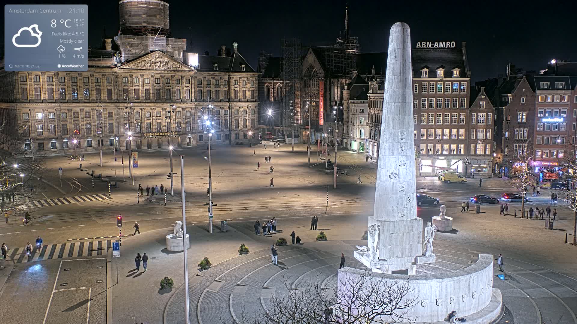 A nighttime, mostly clear view of a large city square with a tall monument at its center, surrounded by buildings and a few pedestrians.