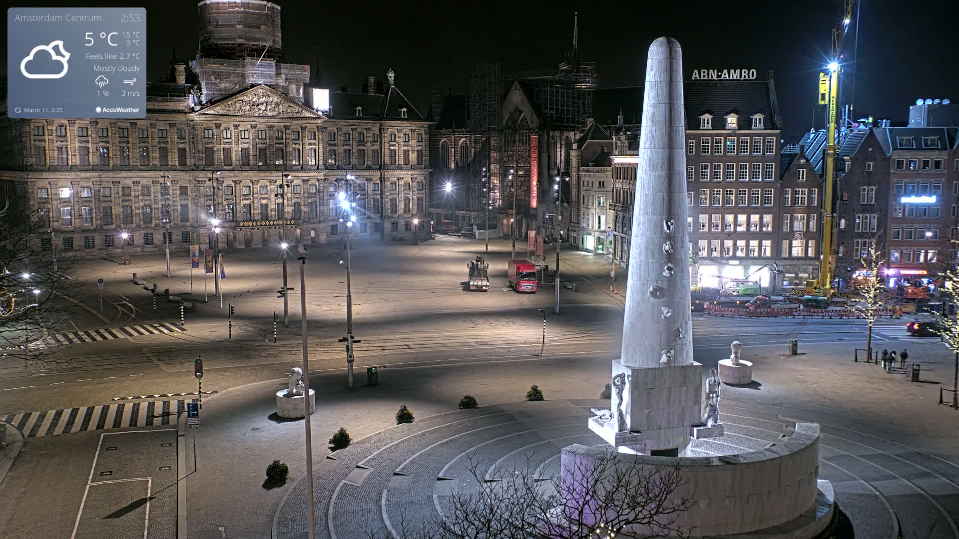 A mostly cloudy night scene shows a large, empty plaza with a tall monument at its center, surrounded by buildings, including a large palace.
