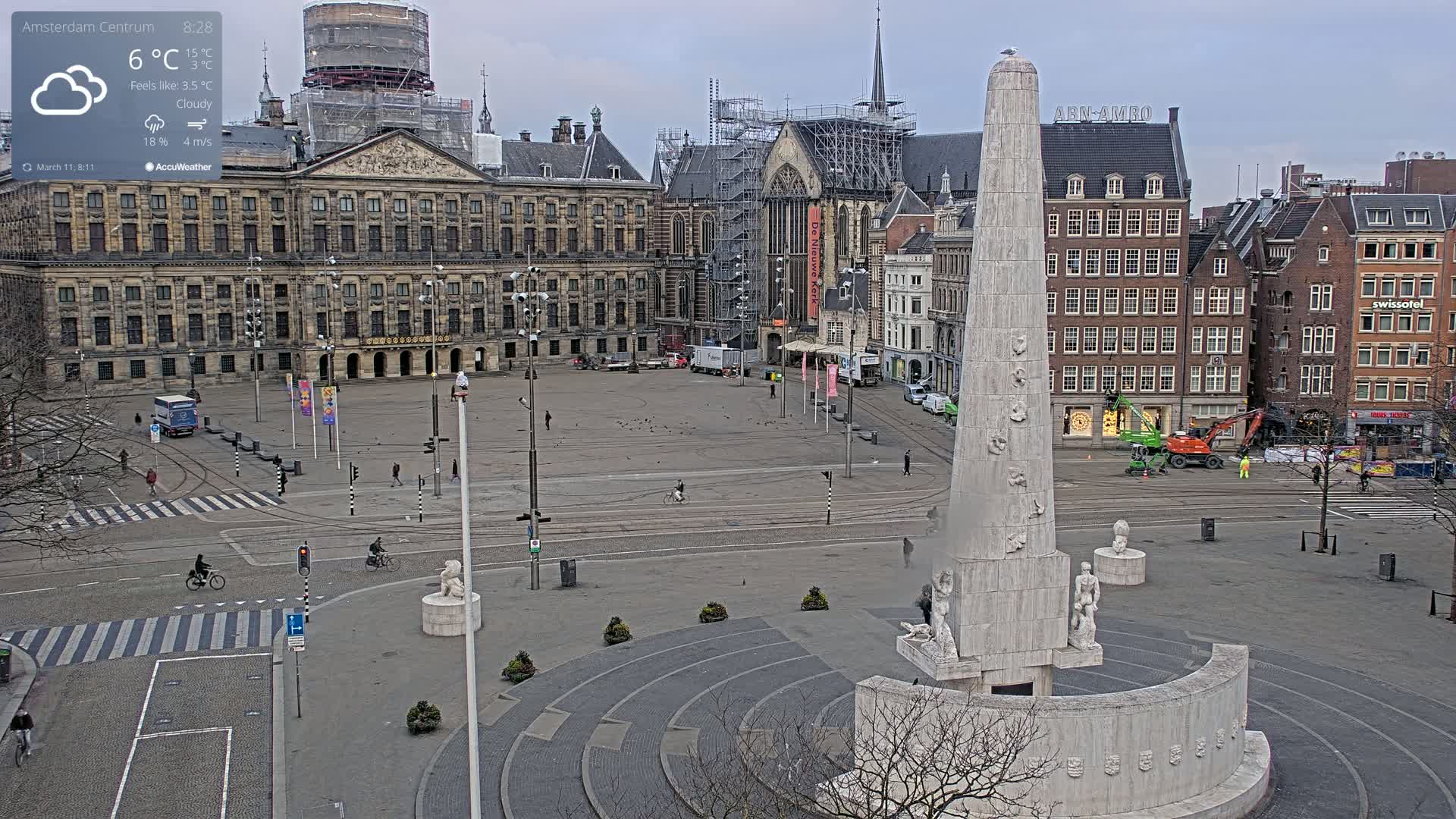 A mostly empty city square on a cloudy day, centered on a large monument, with several buildings and a few people and bicycles visible.