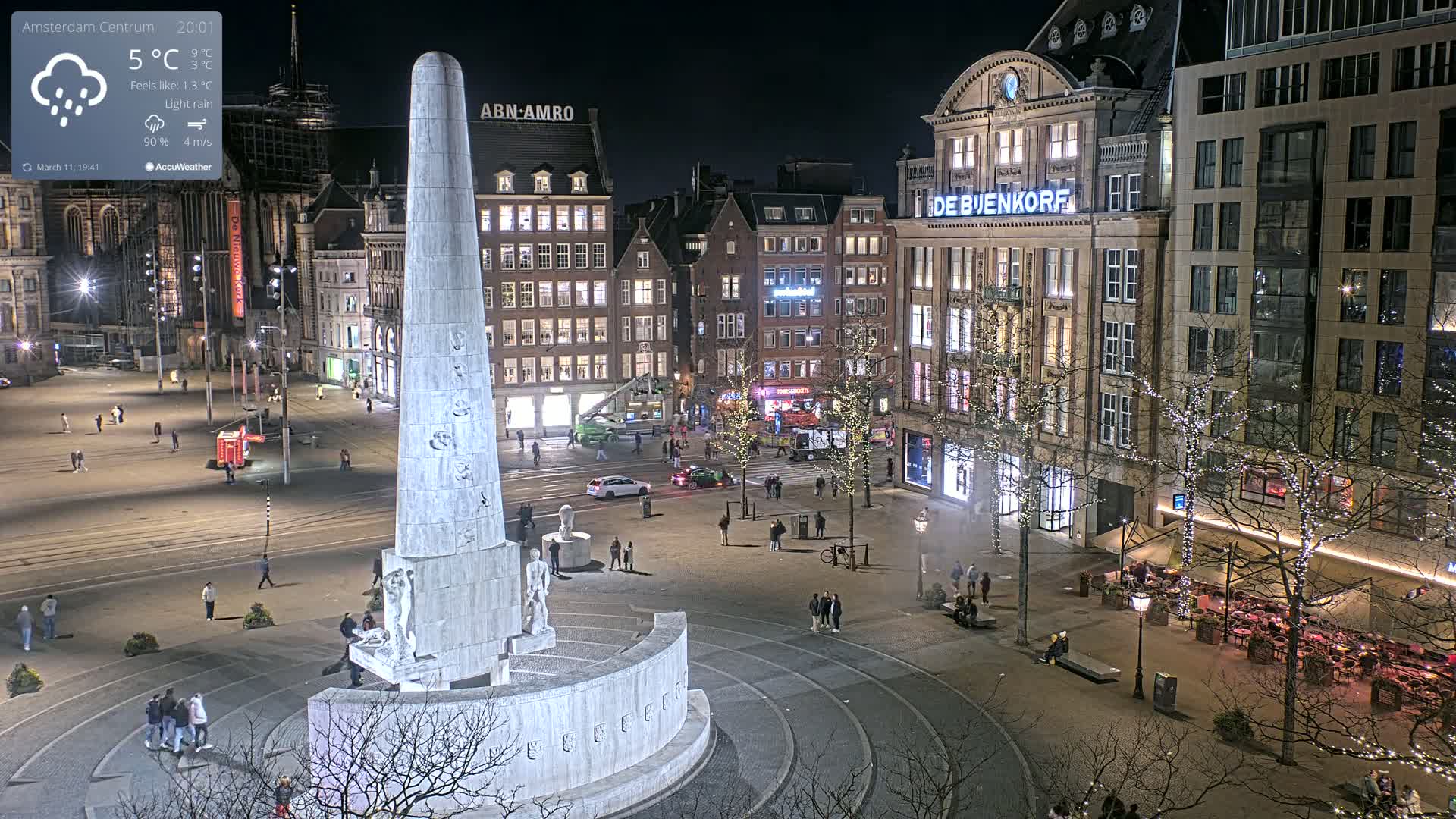 A nighttime, lightly raining view of a city square, centered on a tall monument, surrounded by buildings with illuminated windows and streetlights, and sparsely populated with people.