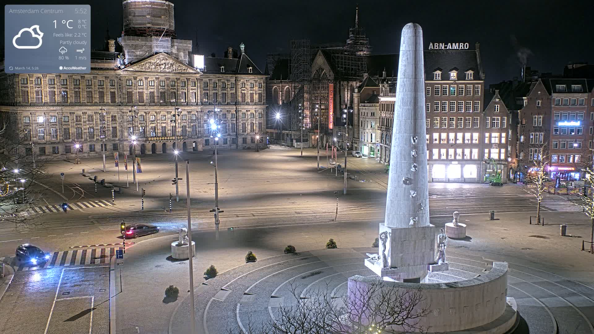 A mostly empty city square at night, under partly cloudy skies, features a large monument and a palace in the background.