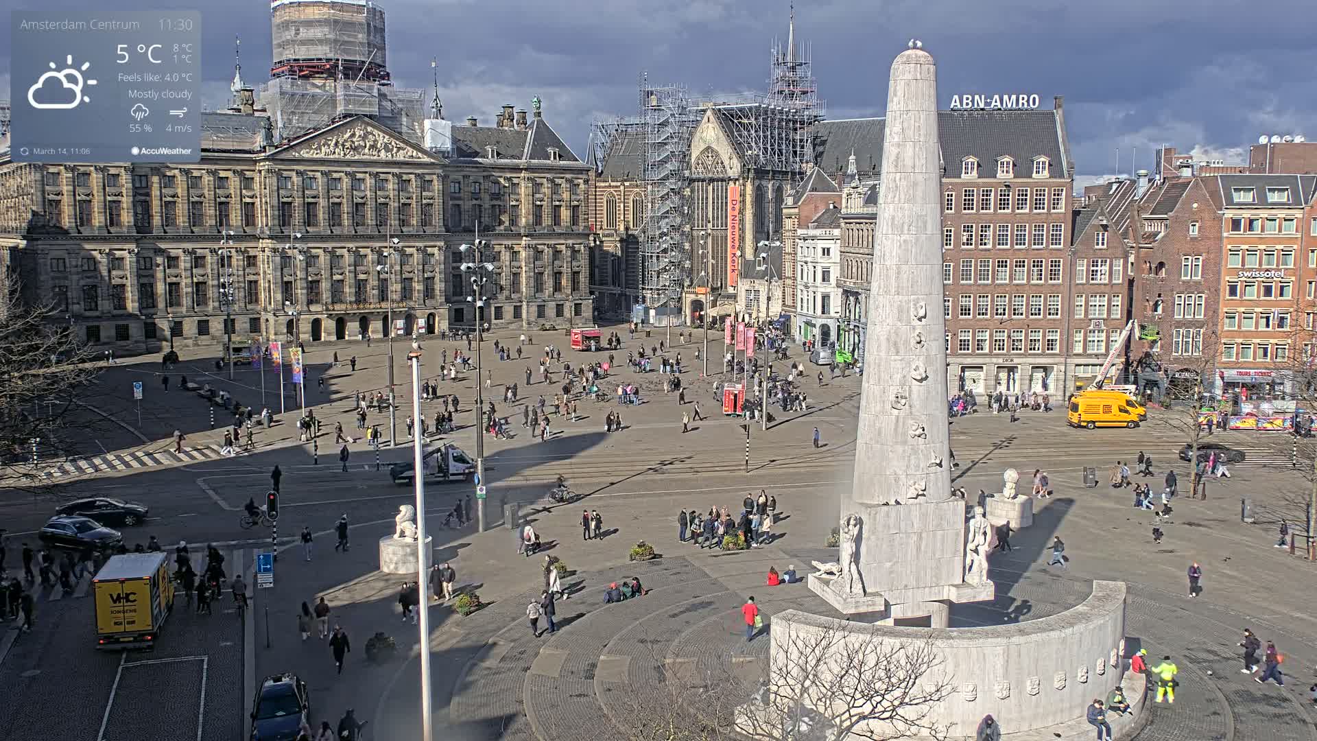 A mostly cloudy day in a large city square with a tall monument at its center, surrounded by buildings and people.