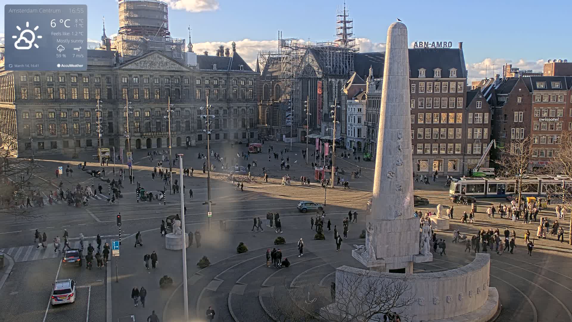 A mostly sunny city square with a tall monument in the center, surrounded by buildings, people, and a tram, under a partly cloudy sky.