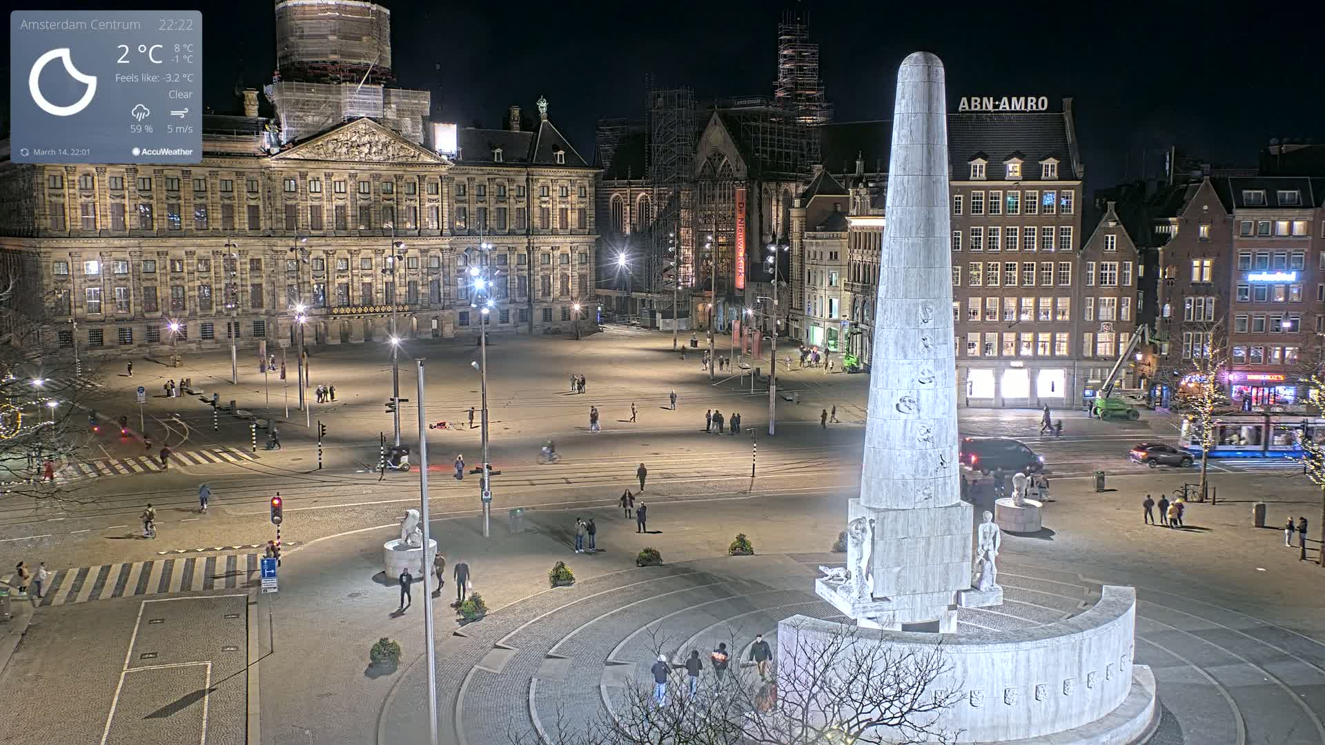 A nighttime, clear view of a large city square with a tall monument at its center, surrounded by buildings and sparsely populated with pedestrians.