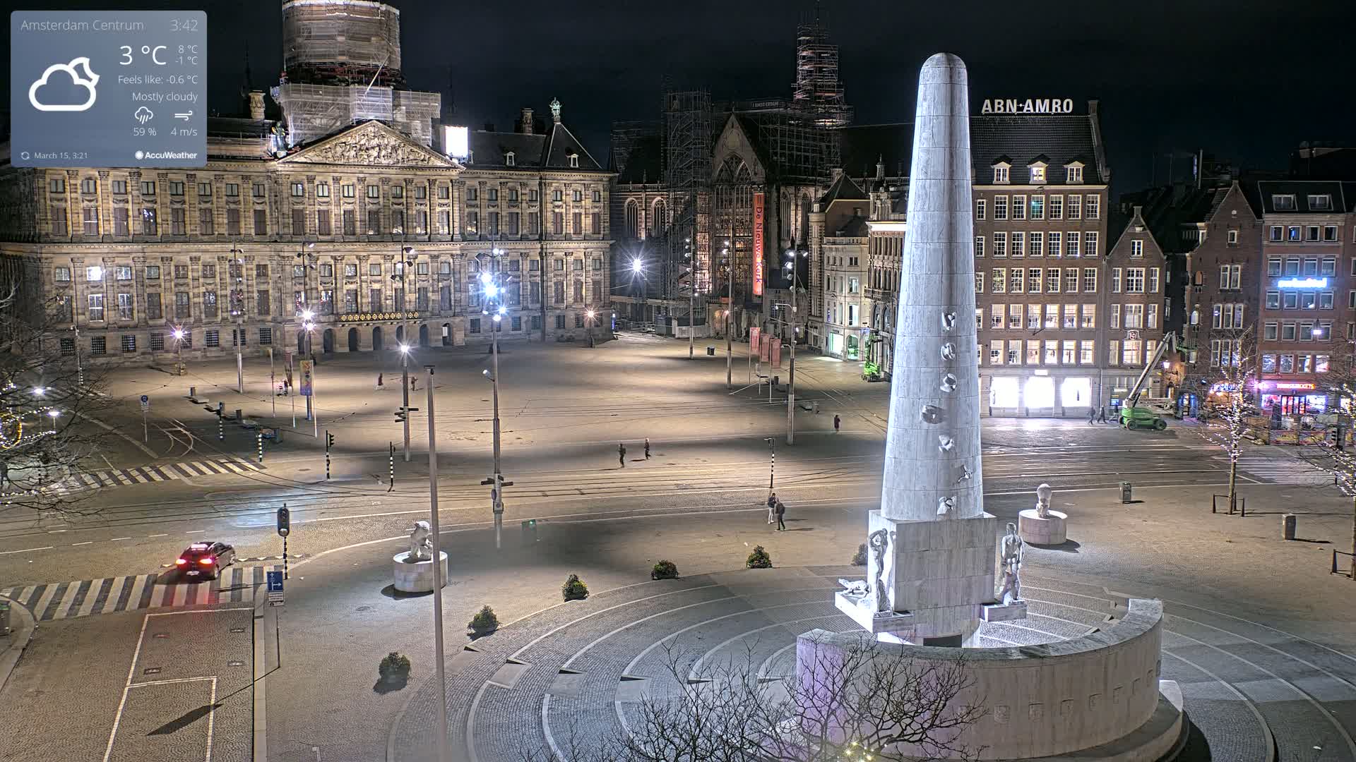 A mostly cloudy night scene shows a large, empty plaza with a tall obelisk-like monument at its center, surrounded by illuminated buildings and streets.