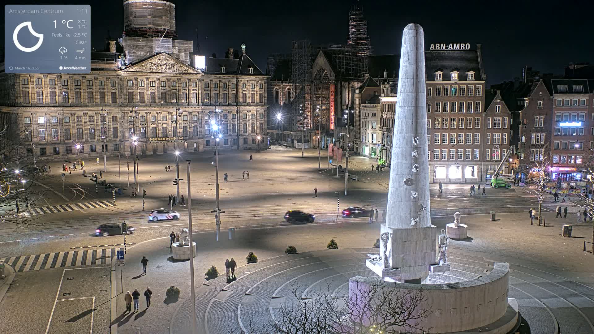 A nighttime, clear view of a large city square with a tall monument at its center, surrounded by historic buildings and a few cars and pedestrians.