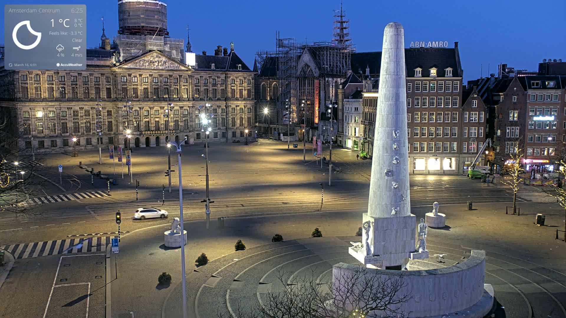 A mostly empty city square at night under a clear sky, illuminated by streetlights, features a tall monument in the foreground and historic buildings surrounding it.