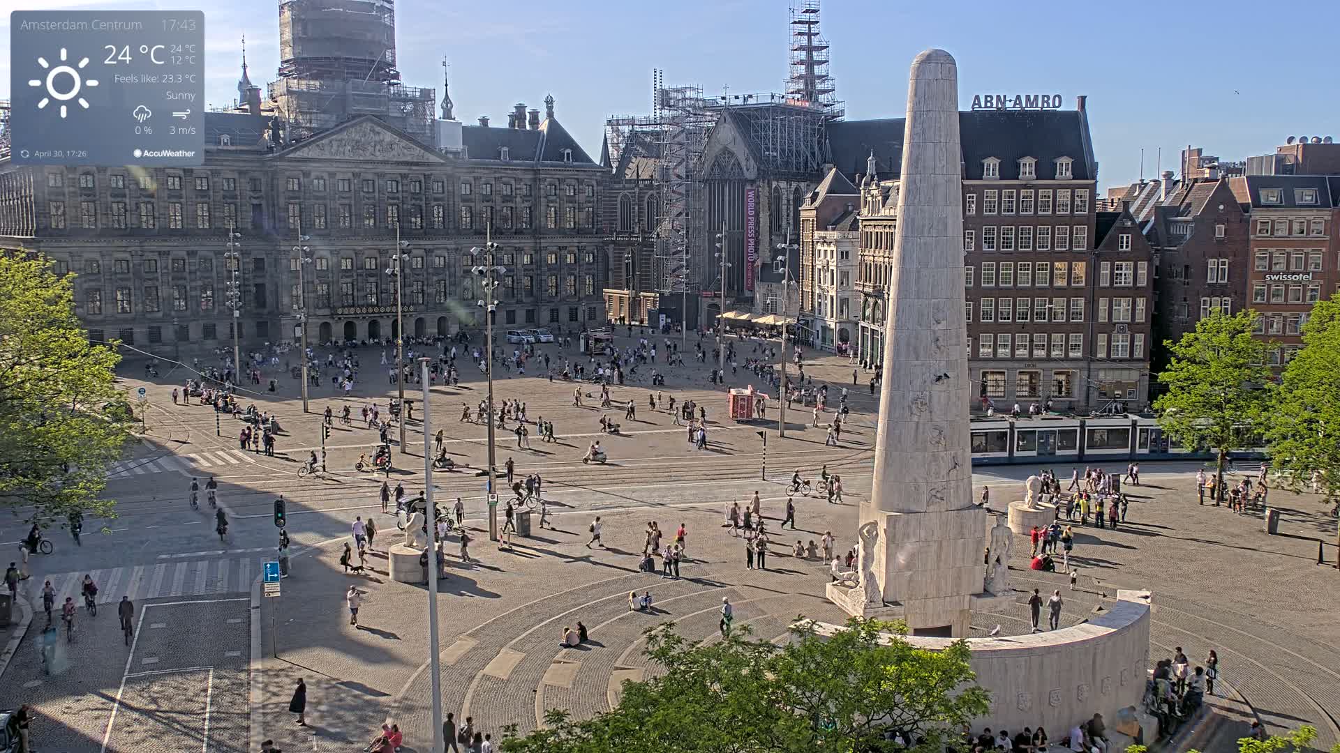 A sunny plaza with a tall monument at its center, surrounded by historical buildings and people, under a clear sky.