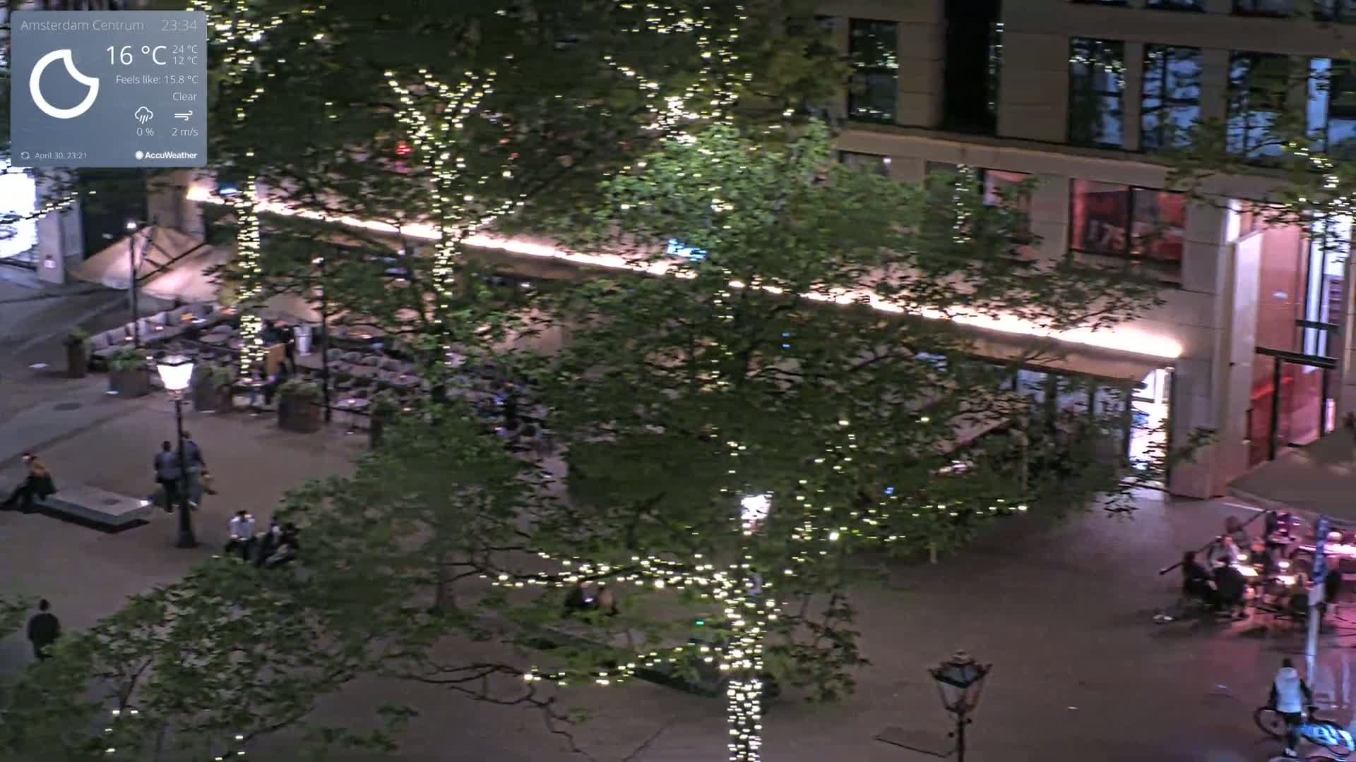 An aerial nighttime view shows a city street with people walking and sitting at outdoor cafes beneath trees adorned with string lights, under clear skies.