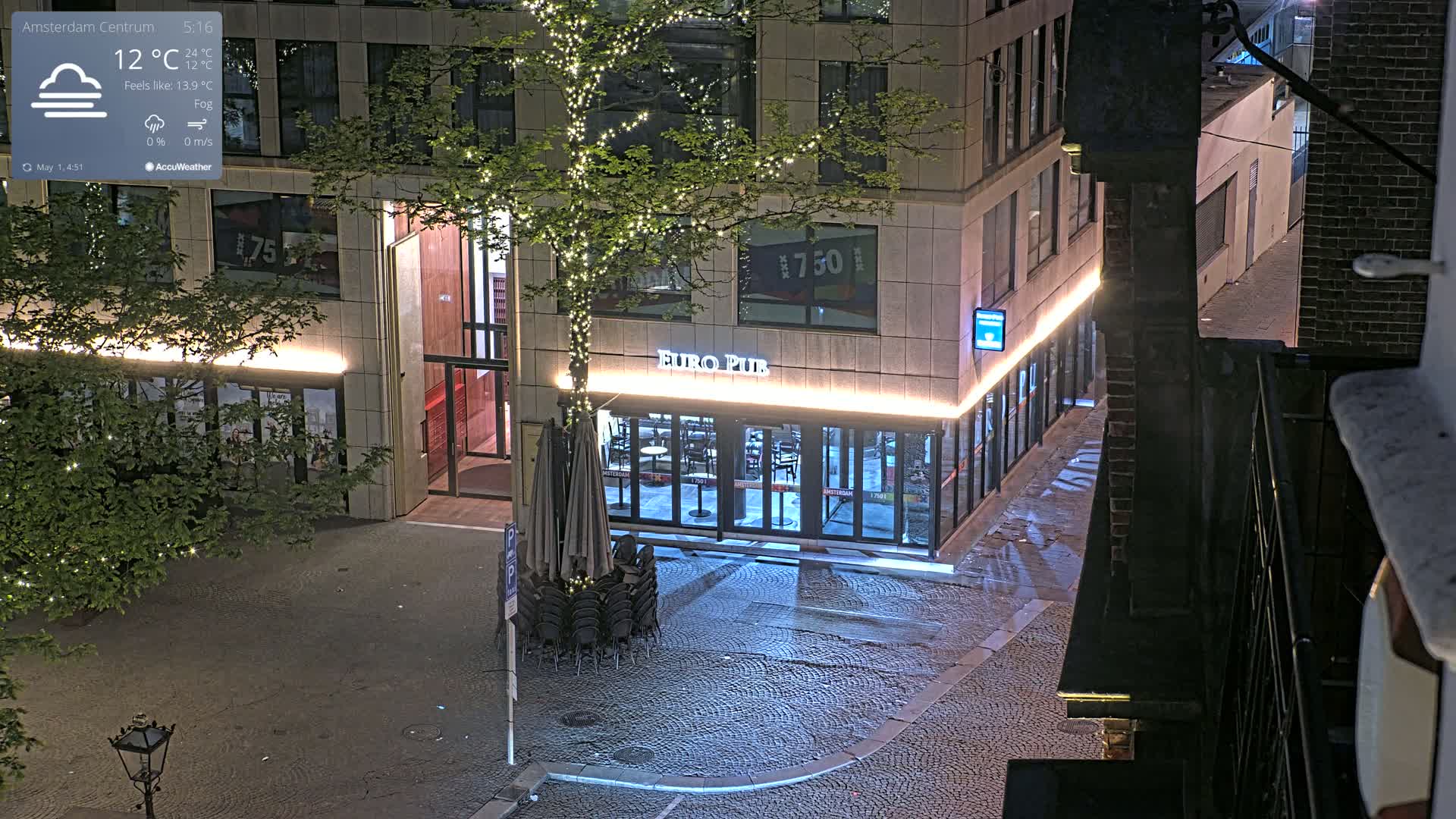 A nighttime, foggy street scene shows a Euro Pub with its chairs stacked outside, next to a tree with string lights, in front of a modern building.