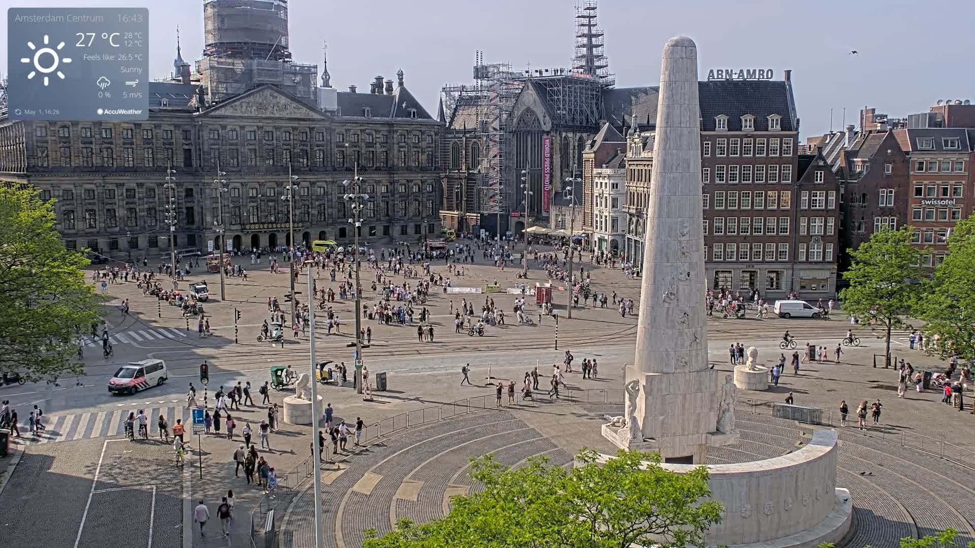 A sunny, 27°C day in a large European square, featuring a monument, numerous pedestrians, and buildings undergoing construction.