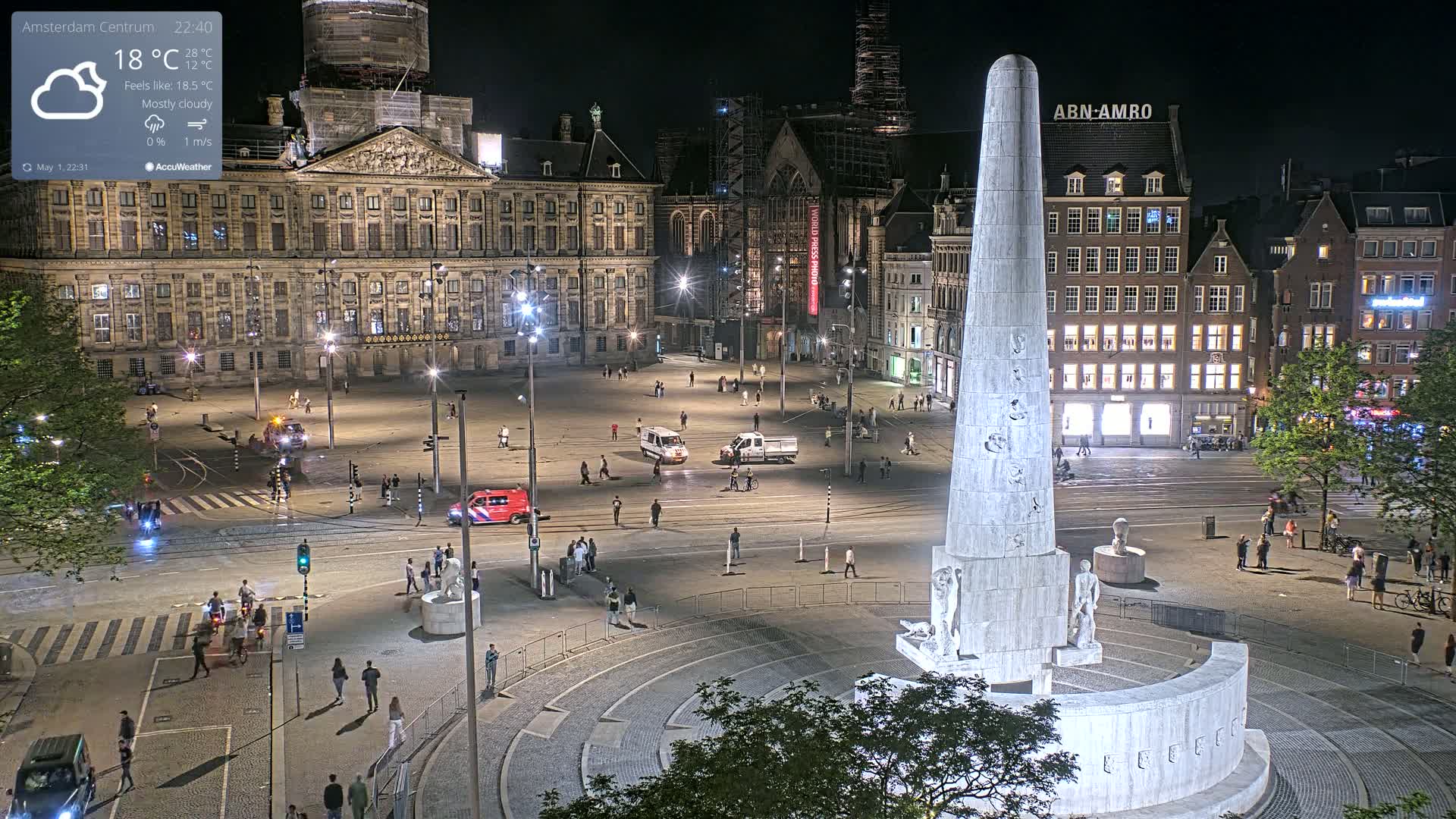 A nighttime, mostly cloudy view of a large, mostly empty square with a tall monument at its center, surrounded by buildings and a few pedestrians.