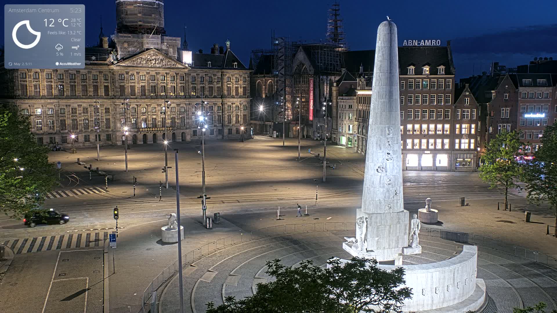 A nighttime, clear view of a city square with a tall monument at its center, surrounded by buildings, including a large palace and modern structures.