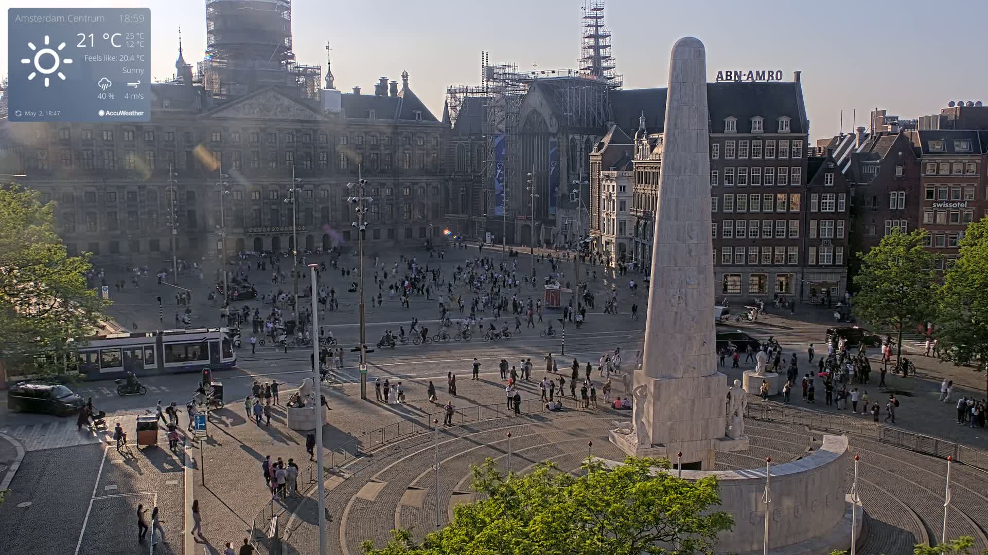 A sunny, bustling city square is viewed from above, centered on a tall monument surrounded by people, with trams and buildings visible in the background.