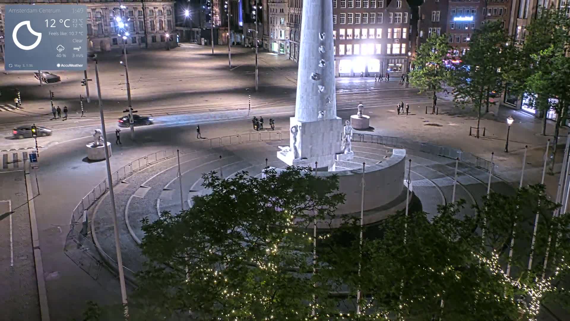 A nighttime, clear view of a city square with a tall monument at its center, surrounded by sparsely populated streets and buildings.