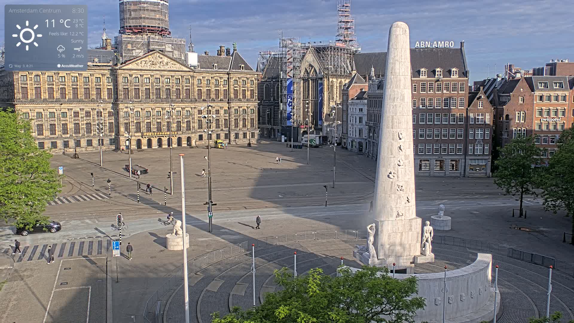 A sunny, mostly empty city square features a tall monument surrounded by classical buildings, one of which is under construction.
