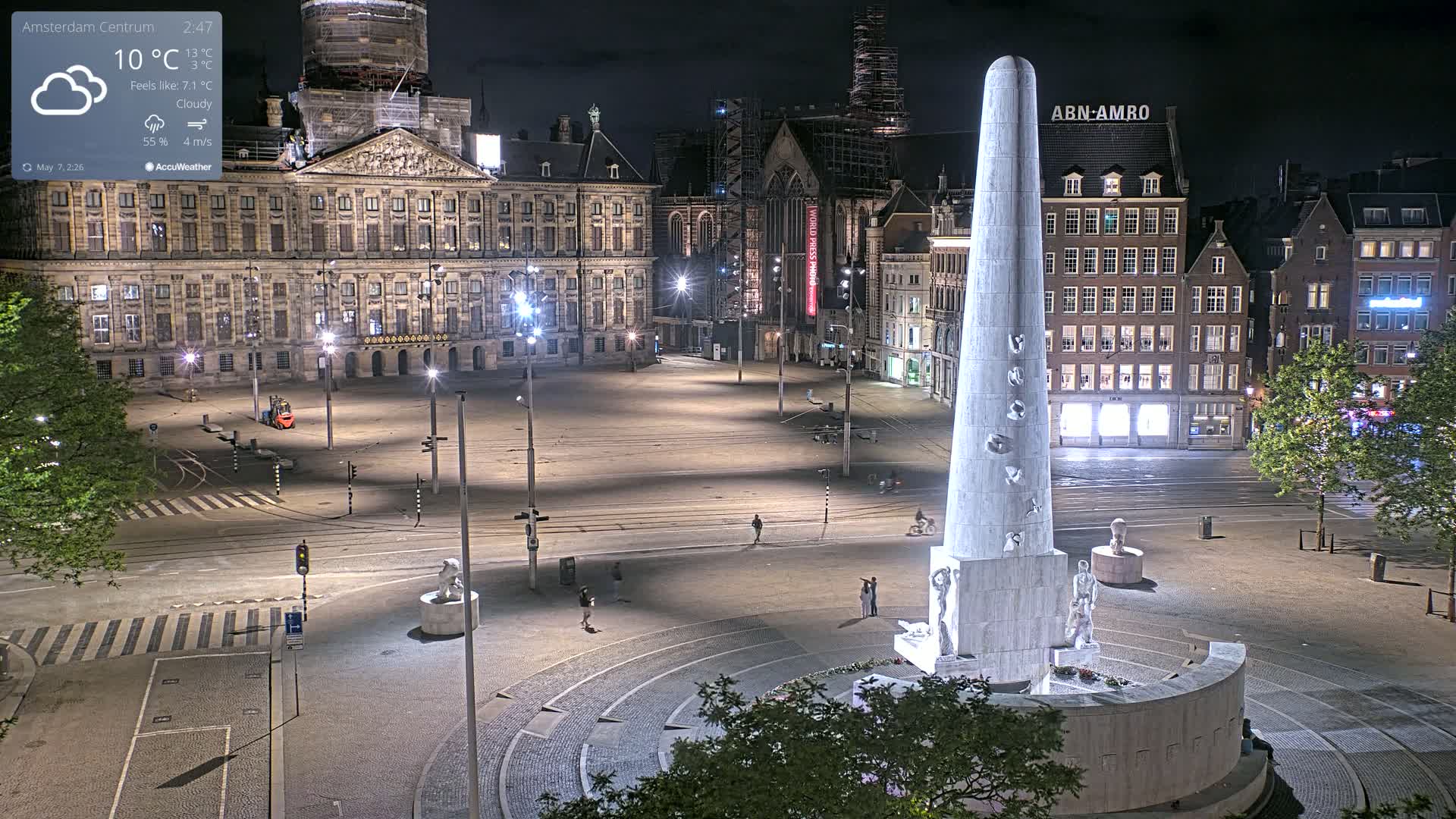 A nighttime, cloudy view of a large, mostly empty city square with a tall monument at its center, surrounded by buildings illuminated by streetlights.