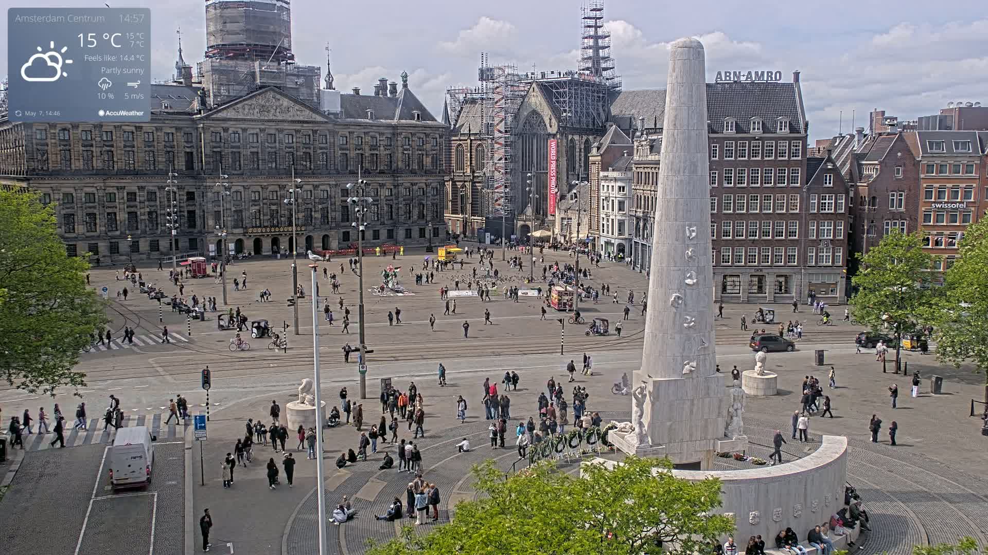 A partly sunny city square with a large monument in the center, surrounded by historical buildings and many people.