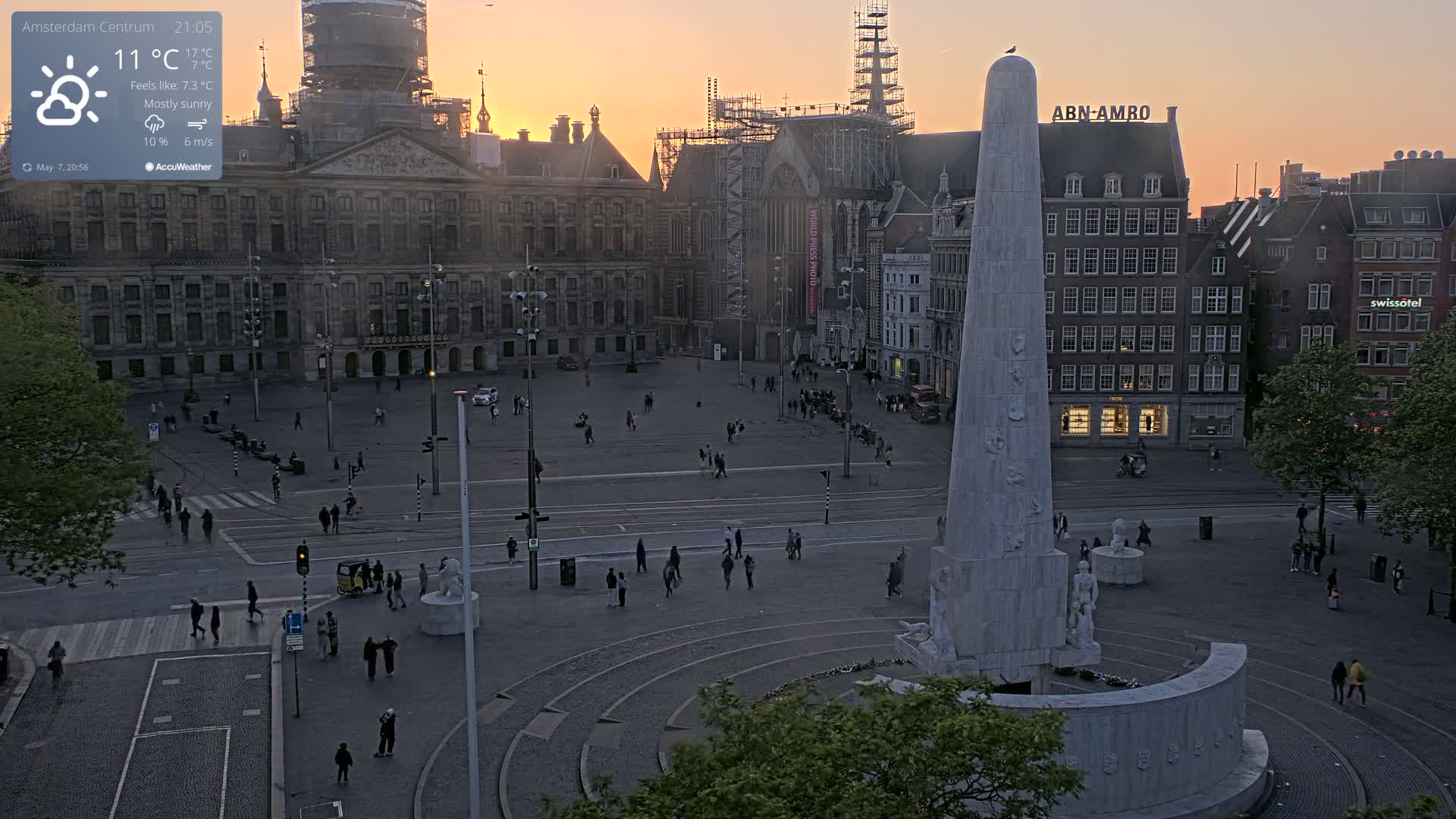 A mostly sunny, cool evening scene shows a large square with a tall monument at its center, surrounded by buildings, with a few people walking around.