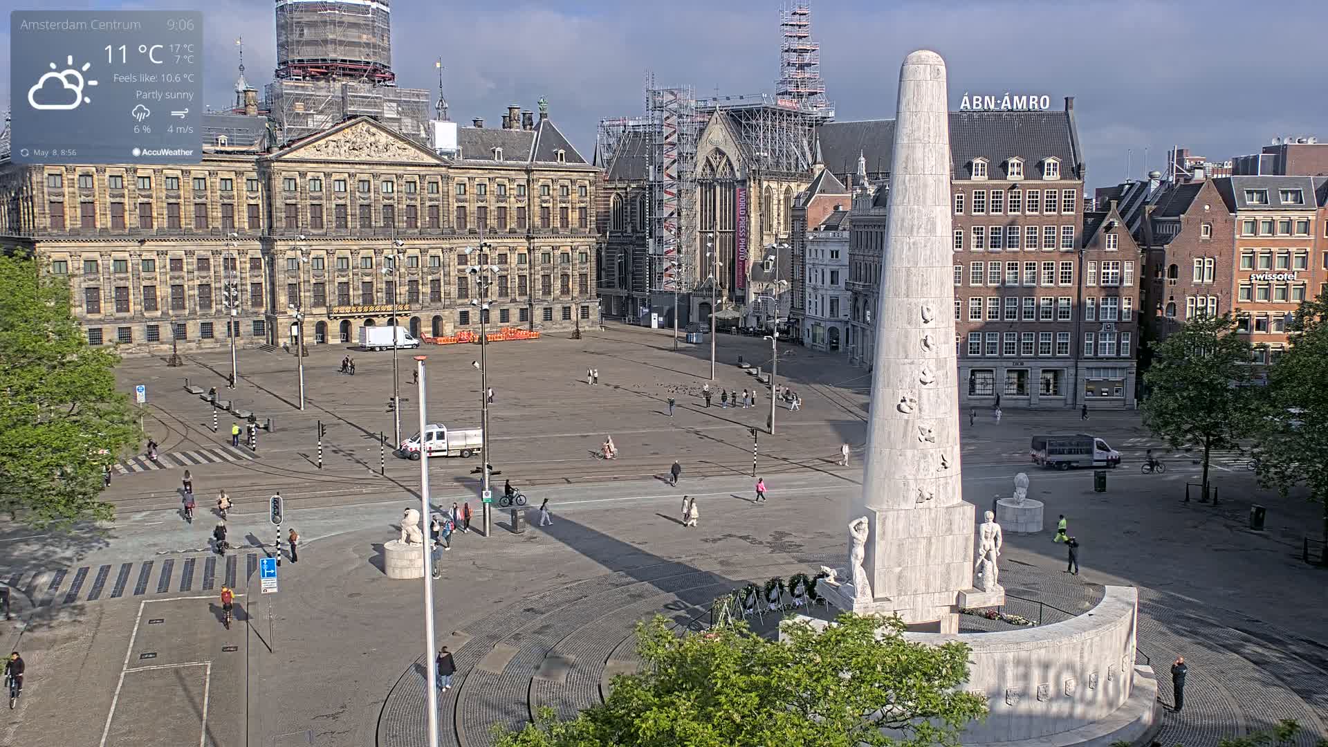 A partly sunny day shows a large city square with a tall monument in the center, surrounded by historical buildings and a few pedestrians.