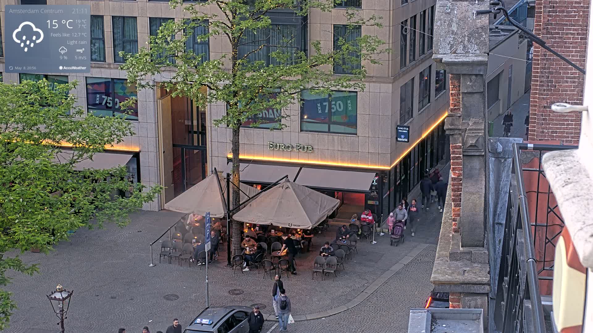 An outdoor cafe with patrons sitting under umbrellas in light rain, situated next to a building with illuminated signage.