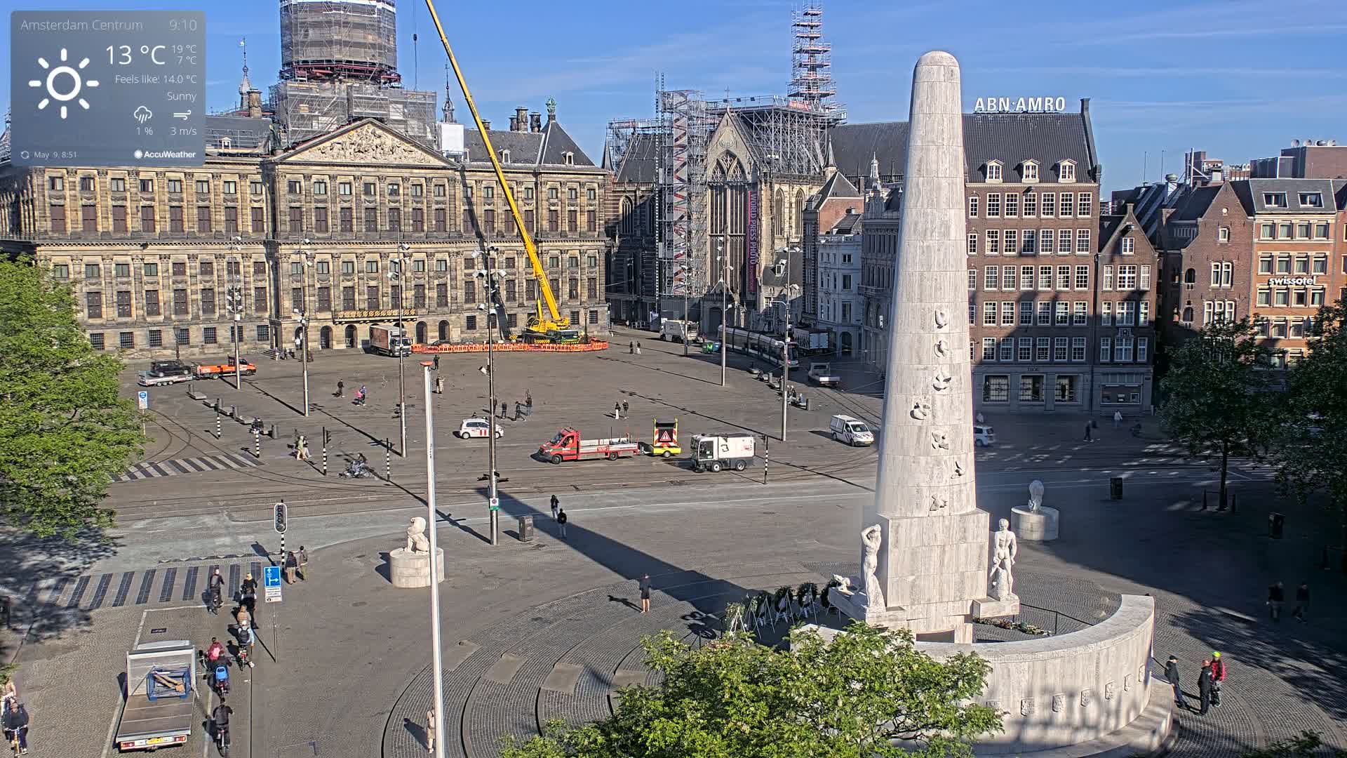 A sunny city square with a tall monument at its center, surrounded by buildings, including a large palace under construction, with several people and vehicles visible.